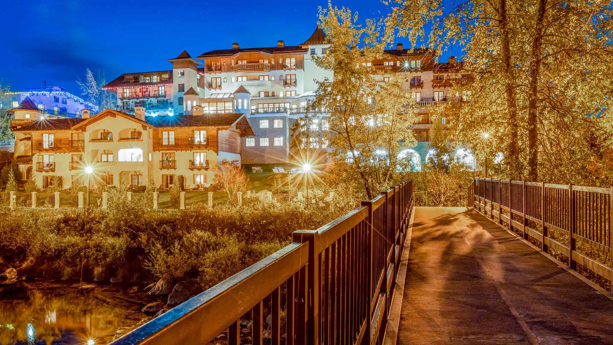 A Bavarian-style alpine village in Leavenworth, Washington is brightly lit against a night sky, with a pedestrian bridge in the foreground leading toward the cozy, chalet-like buildings.