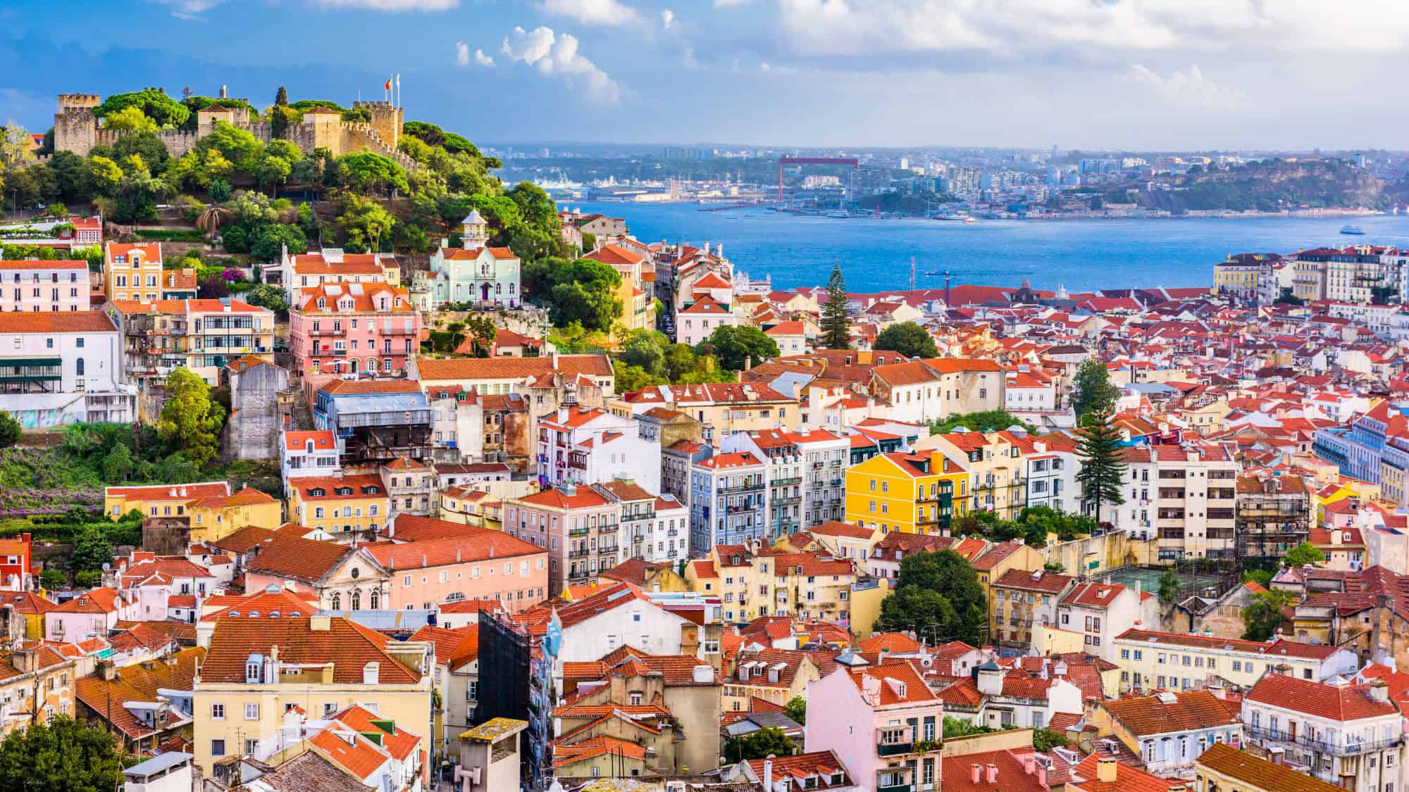 A colorful view of Lisbon, Portugal, featuring a dense cluster of pastel-colored buildings with red rooftops leading up to the historic São Jorge Castle. The Tagus River is visible in the background under a partly cloudy sky.