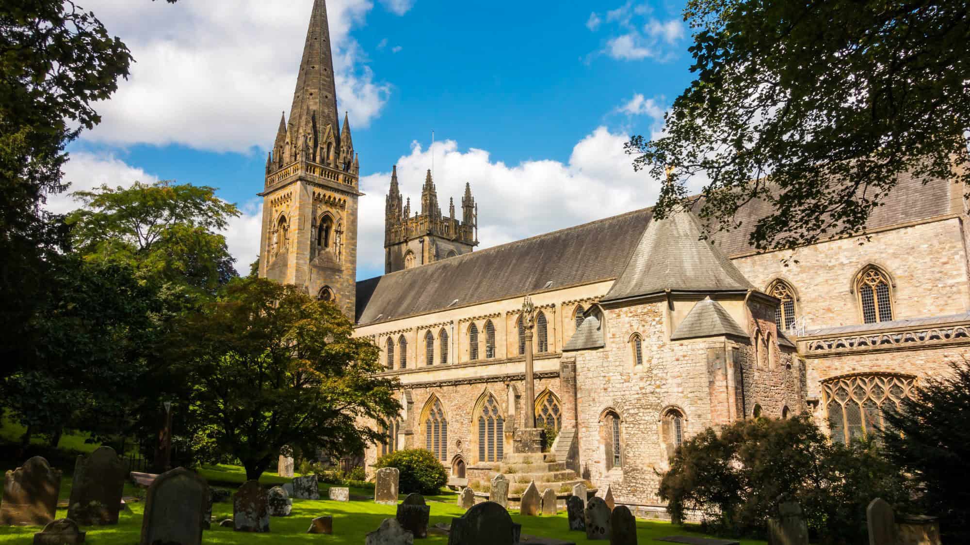 Gothic-style Llandaff Cathedral seen from its churchyard, with tall spires and arched stained-glass windows surrounded by gravestones and trees under a partly cloudy sky.