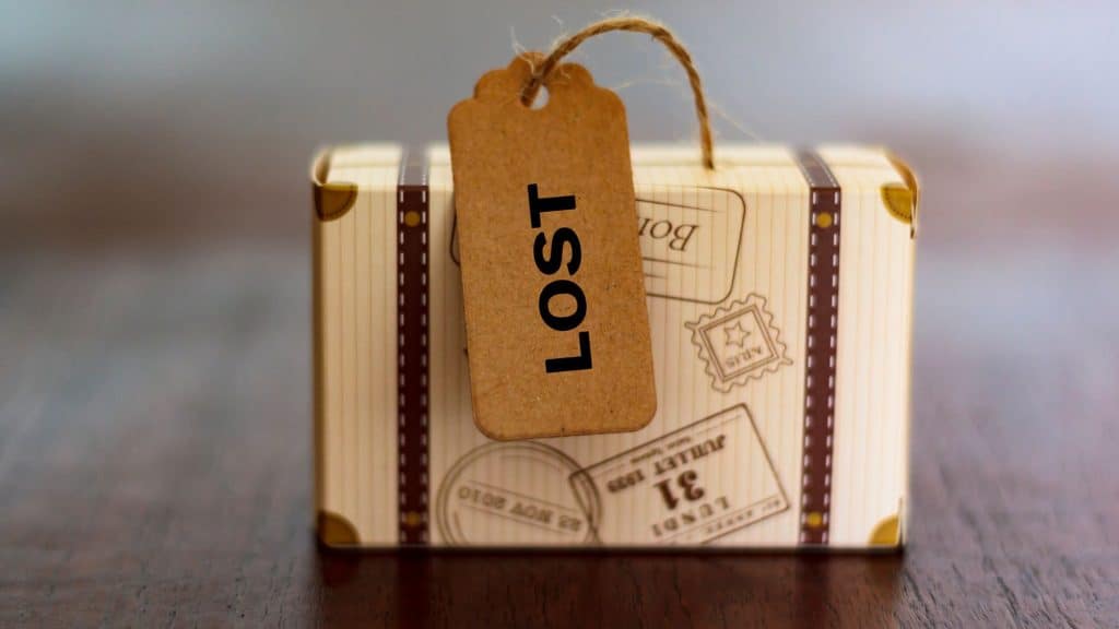 A small decorative suitcase with a vintage travel design sits on a wooden surface, featuring a luggage tag labeled "LOST" in bold black letters.