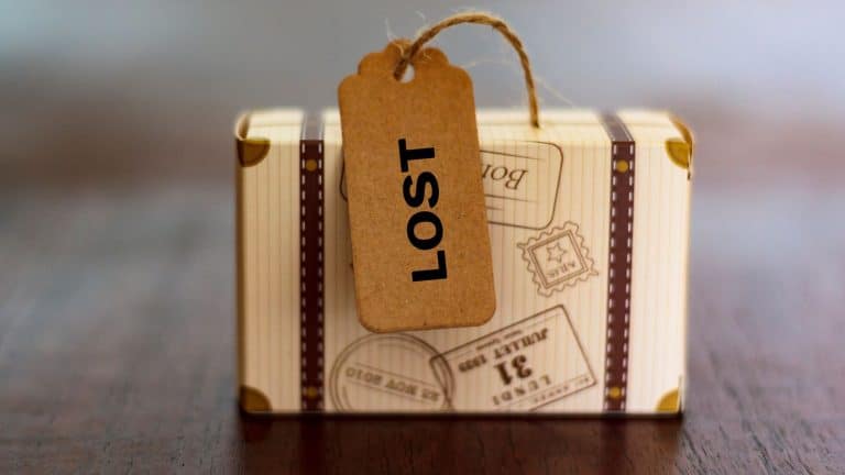 A small decorative suitcase with a vintage travel design sits on a wooden surface, featuring a luggage tag labeled "LOST" in bold black letters.