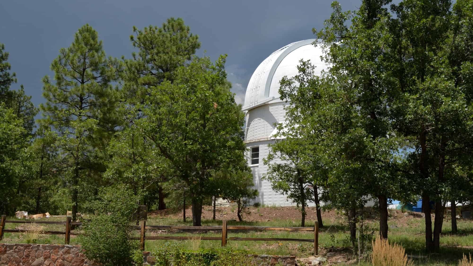 A white domed observatory peeks through lush green trees in a forest setting, with a wooden fence in the foreground. The sky is cloudy, giving a dramatic contrast to the scene.