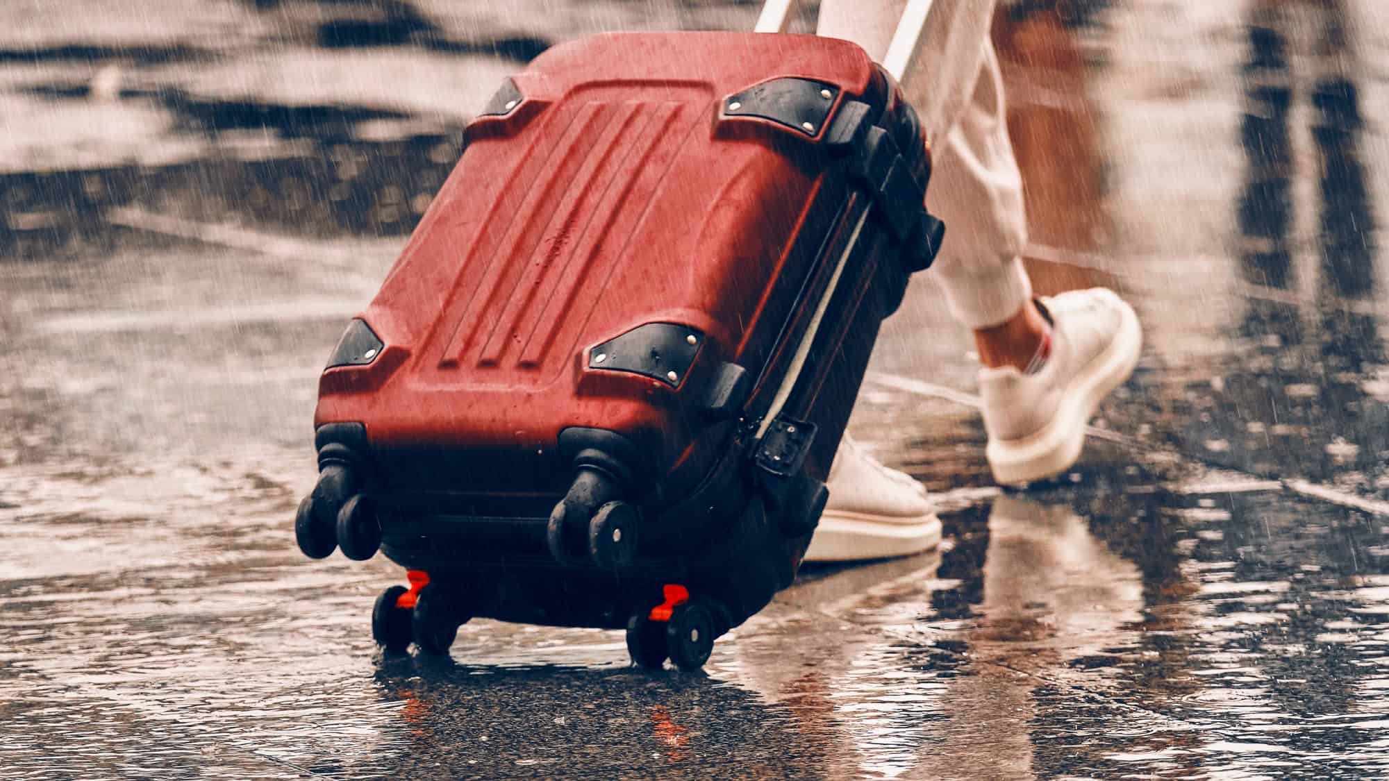 A traveler pulls a wet red suitcase with black trim through a rainy street, wearing white sneakers. Raindrops splash onto the pavement, emphasizing the motion and weather conditions.