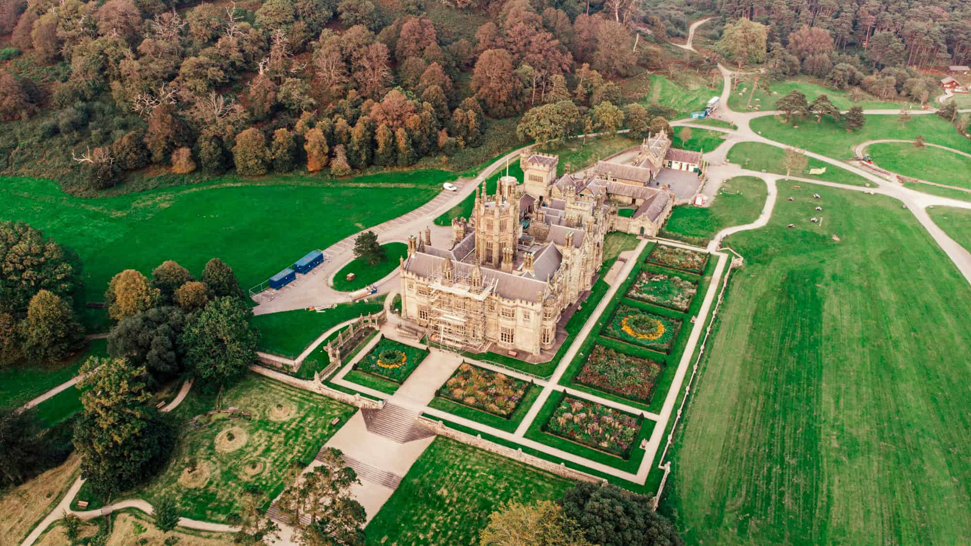 An aerial view of the sprawling Margam Castle estate in Wales, showcasing its ornate Gothic architecture, manicured gardens, and surrounding parkland paths leading into the forest.