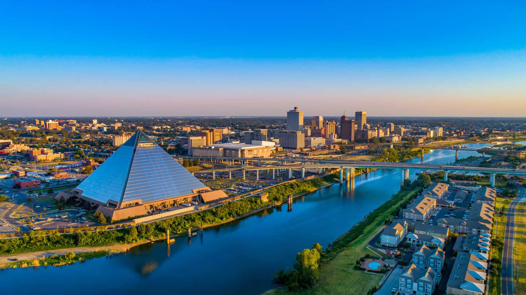 Aerial view of Memphis, Tennessee, showcasing the reflective Memphis Pyramid, the winding Mississippi River with bridges, and a vibrant downtown skyline bathed in warm, golden light.