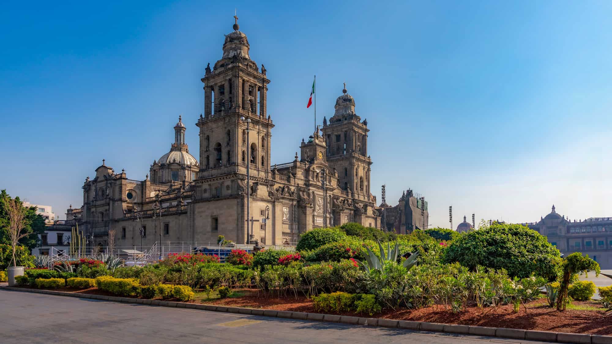 The grand Metropolitan Cathedral of Mexico City with ornate towers and domes, framed by a vibrant garden under a clear blue sky.
