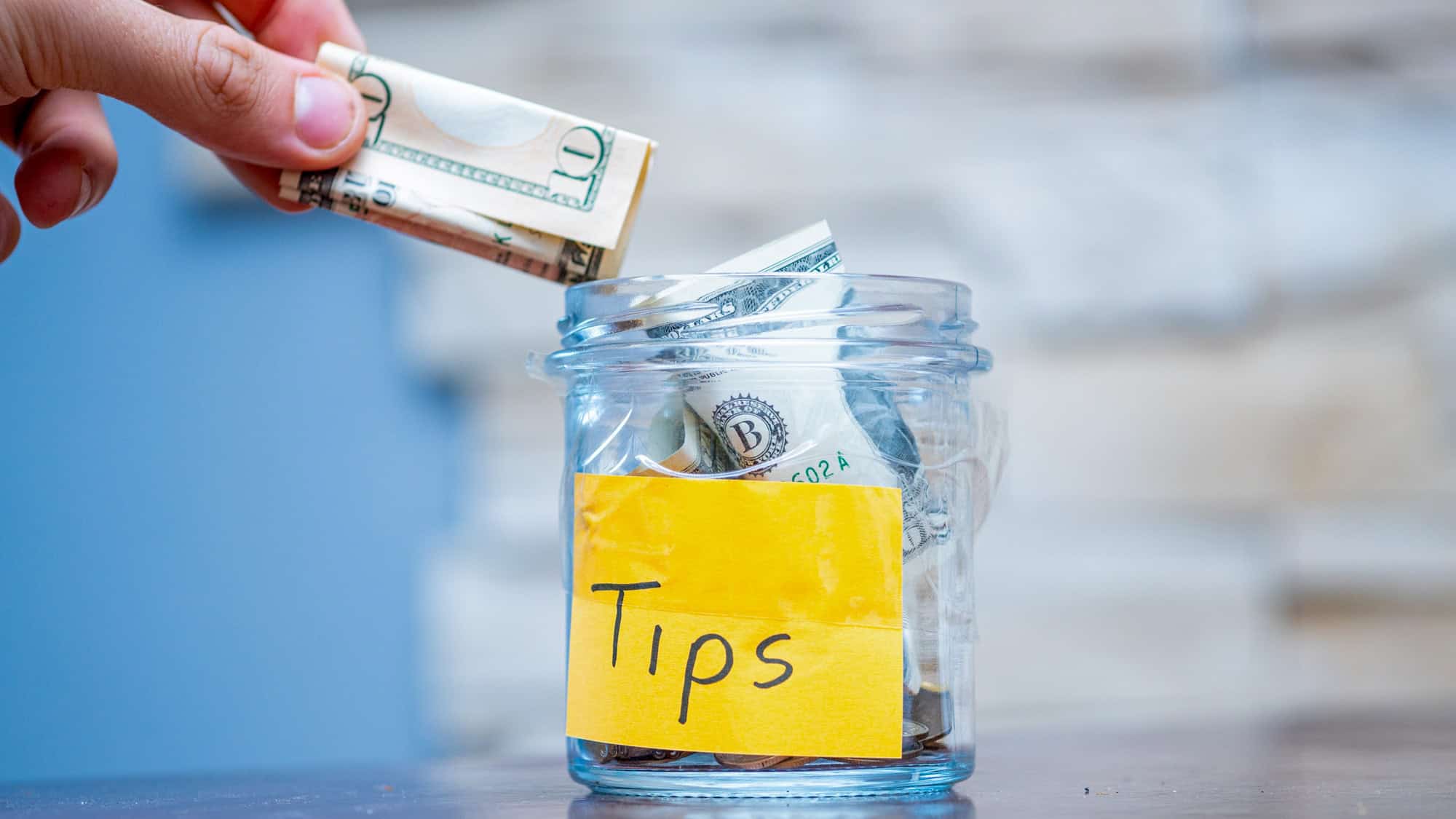 A close-up of a hand placing folded U.S. dollar bills into a clear glass jar labeled "Tips," with additional bills and coins inside, set against a light blue and textured background.