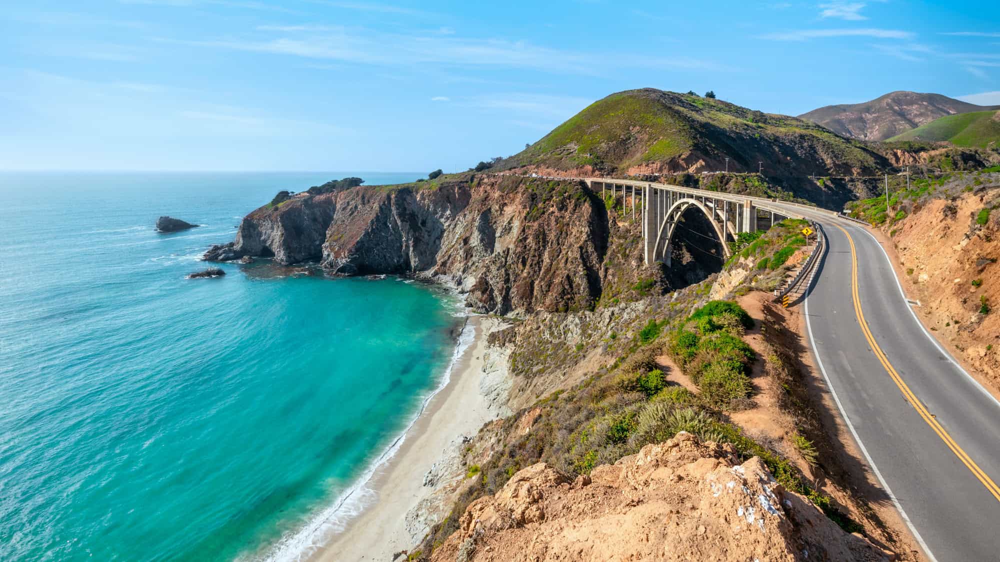 California's iconic Bixby Creek Bridge curves along the rugged Big Sur coastline, with turquoise waters and steep cliffs basking in coastal sunlight.