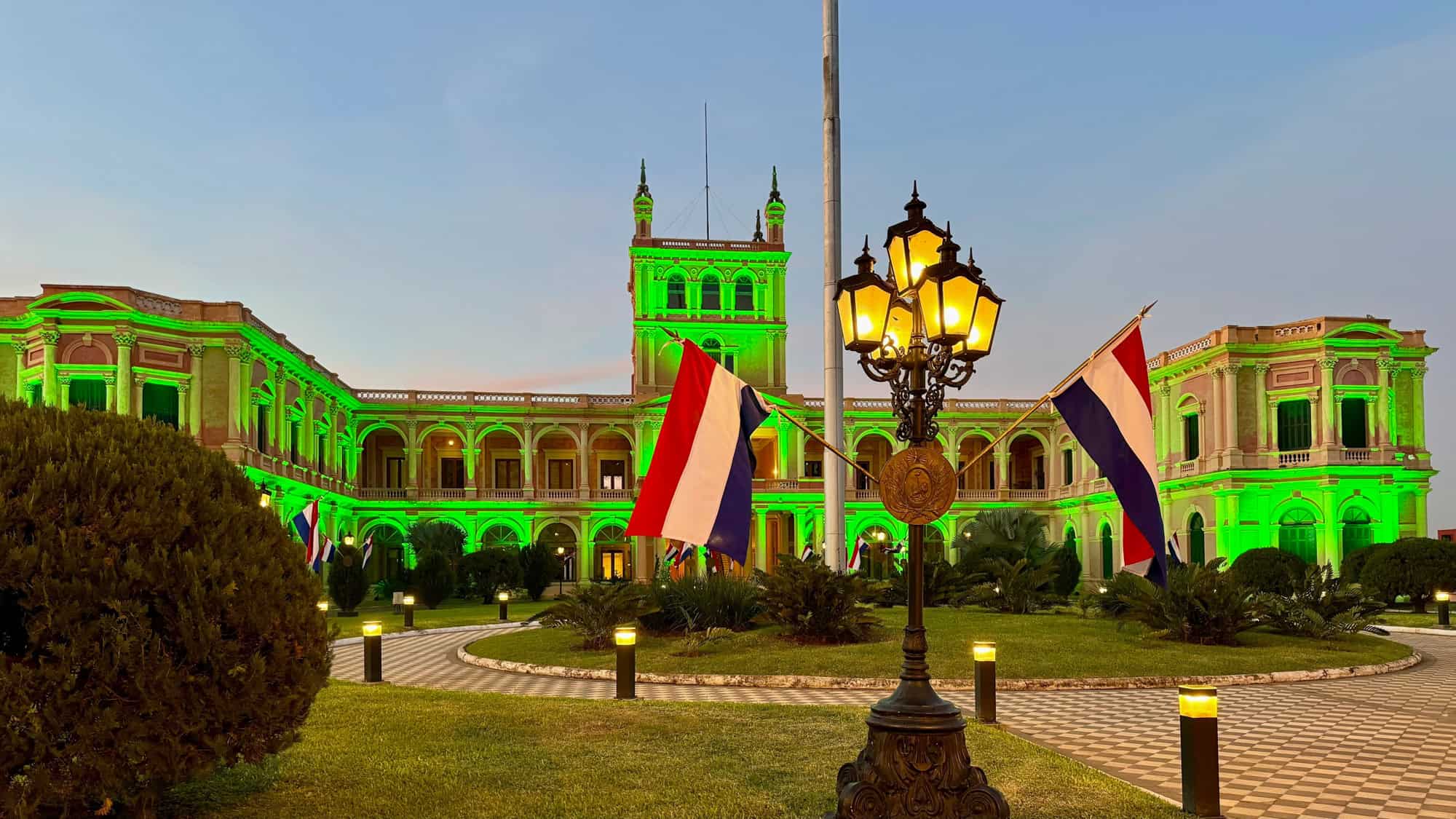Twilight view of the Palacio de los López in Asunción, Paraguay, illuminated with vibrant green lights, surrounded by a manicured garden and Paraguayan flags.