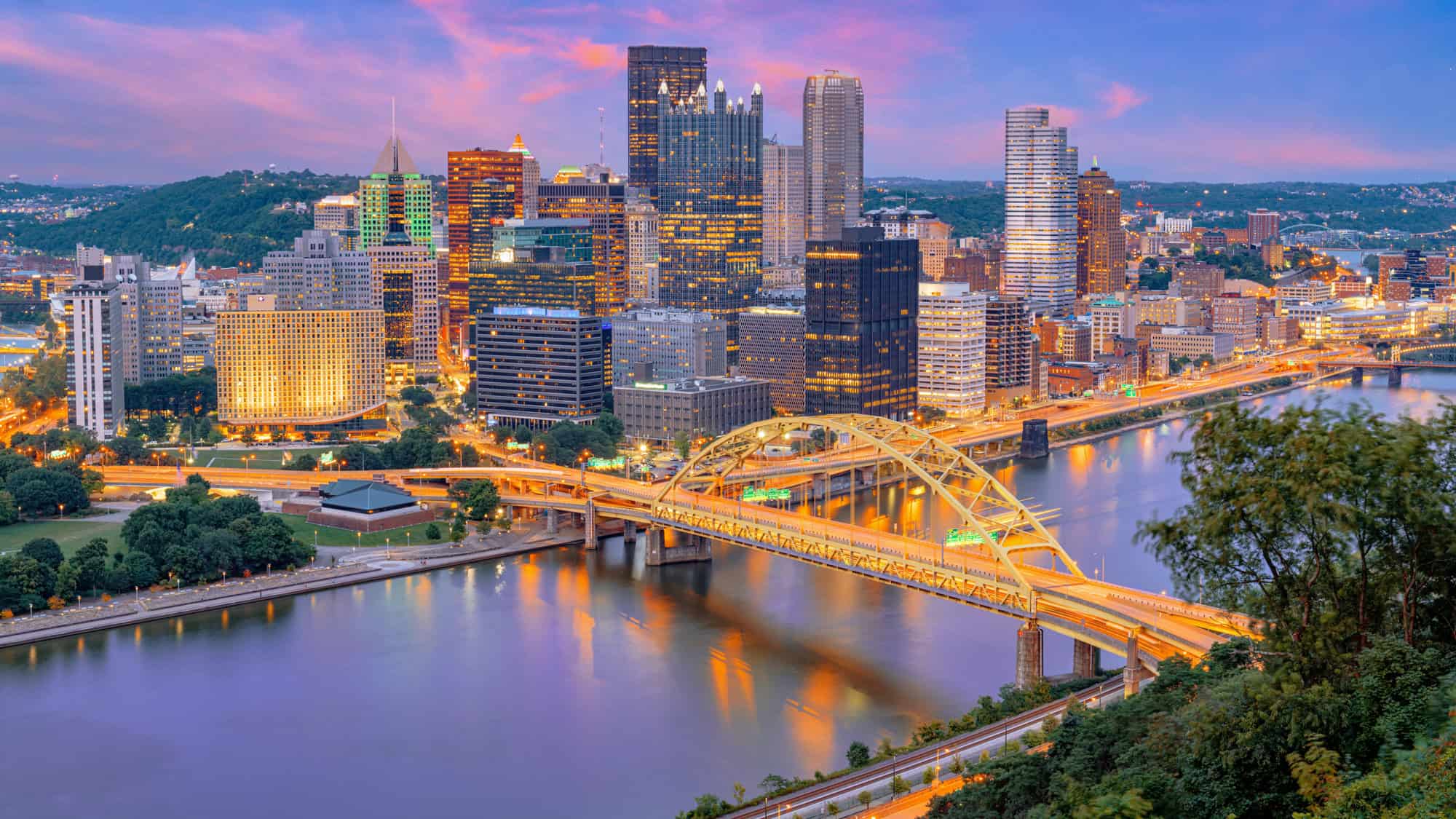 A panoramic view of Pittsburgh's downtown skyline at dusk, featuring the city's signature skyscrapers, bridges, and the meeting point of three rivers with golden reflections on the water.