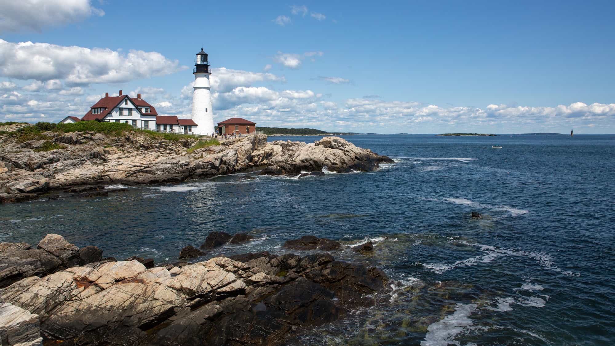 A coastal view featuring the iconic Portland Head Light lighthouse on a rocky promontory, surrounded by the ocean and a bright blue sky.