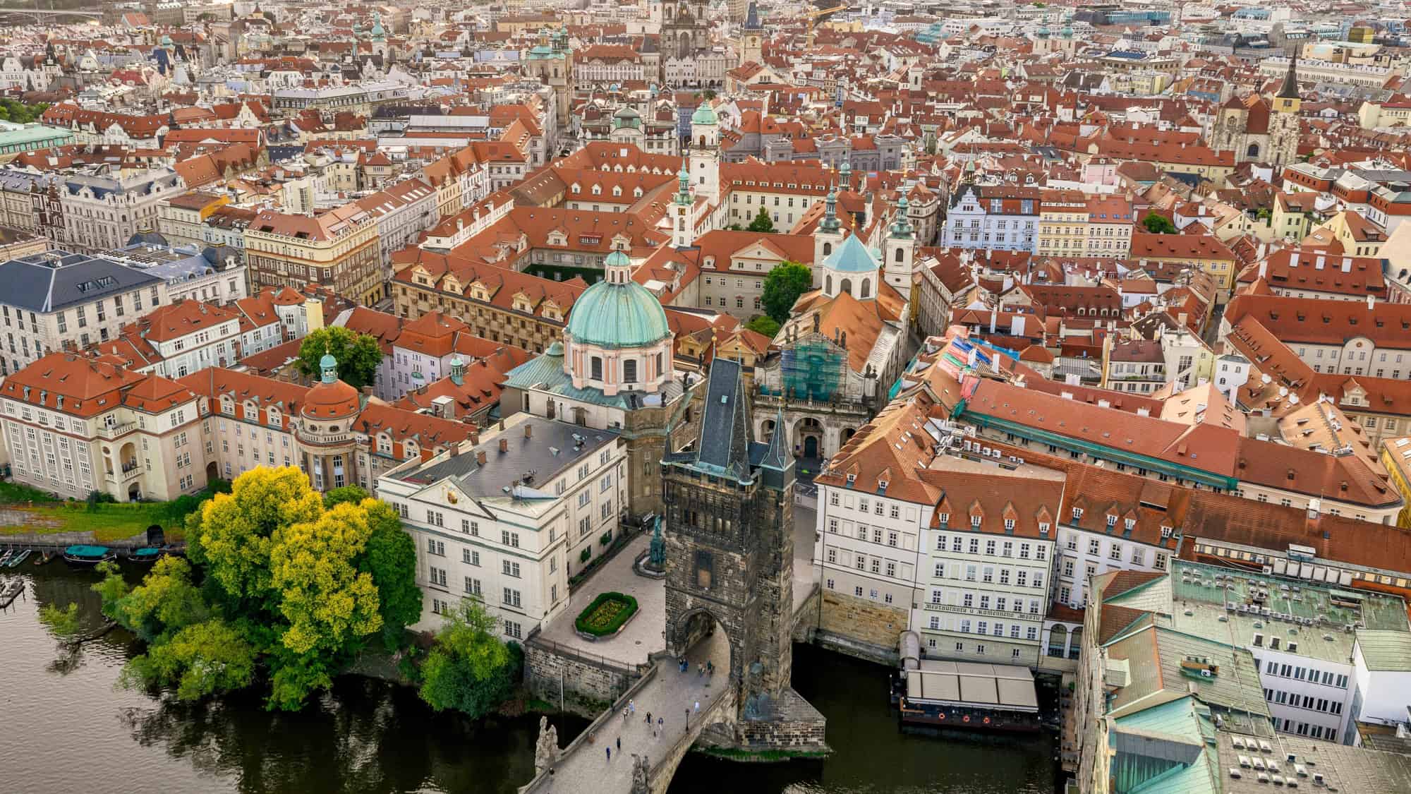 Aerial view of Prague showcasing red-tiled rooftops, the Charles Bridge spanning the Vltava River, and historic landmarks like St. Nicholas Church and the Charles Bridge Tower.