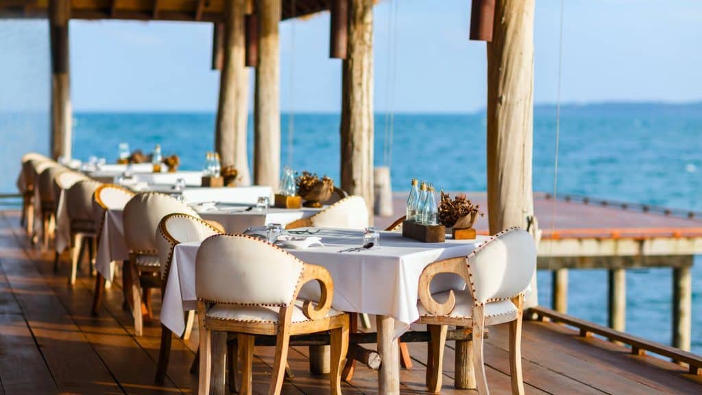 A stunning seaside restaurant with elegantly set tables on a wooden deck overlooking the ocean. The open-air dining space features rustic wooden chairs with cream-colored cushions, offering diners a breathtaking view of the water.
