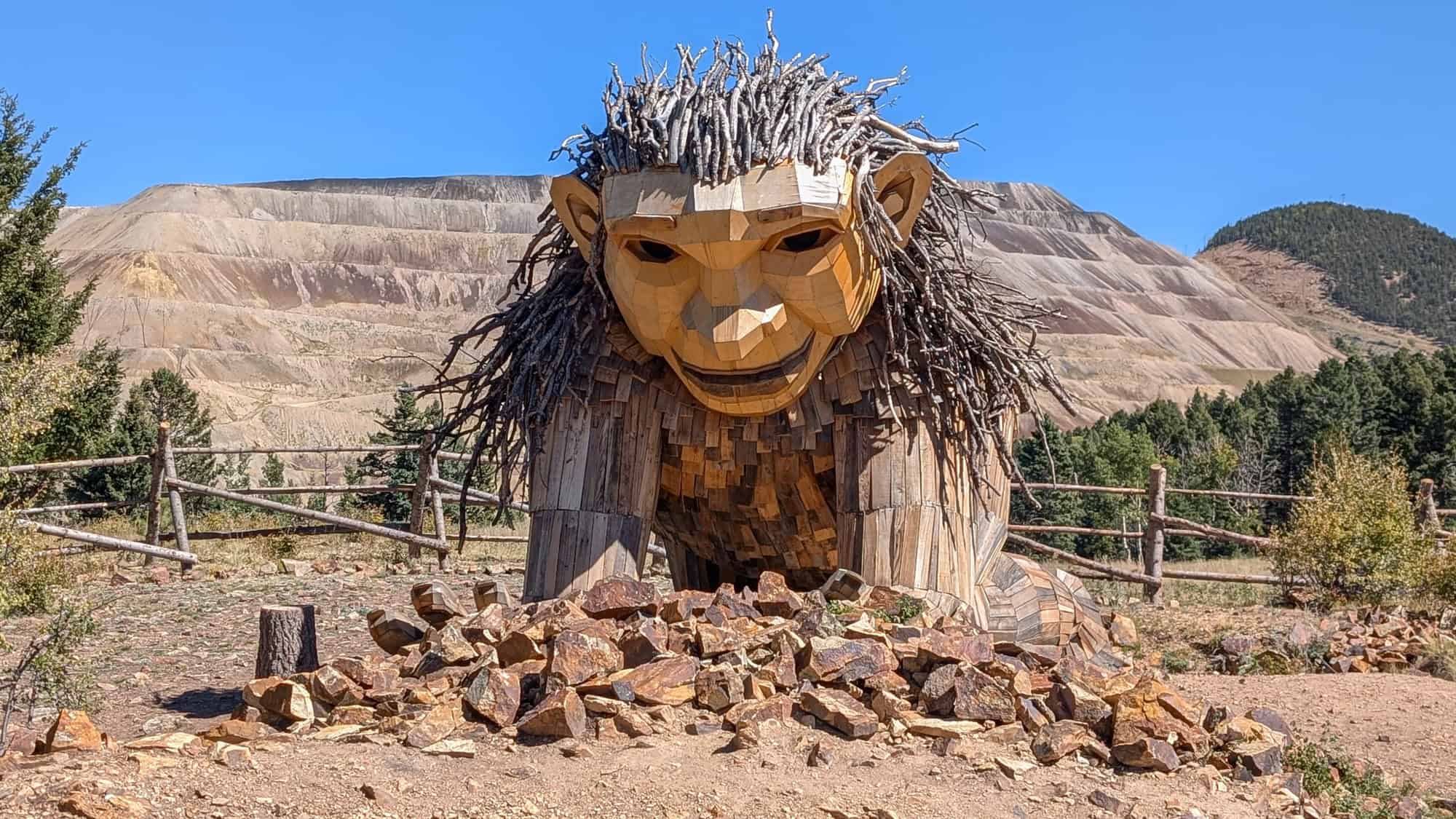 A massive wooden troll sculpture named Rita, made from branches and reclaimed wood, crouches playfully near a pile of rocks. Behind it, a barren mountainside and pine trees create a striking natural backdrop.