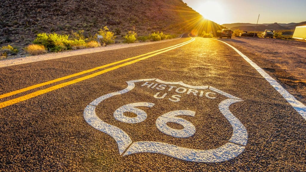 A low-angle view of Historic Route 66 at sunset, featuring the iconic "HISTORIC US 66" shield painted on the asphalt, with golden light illuminating the road, arid hills, and vintage cars parked along the shoulder.
