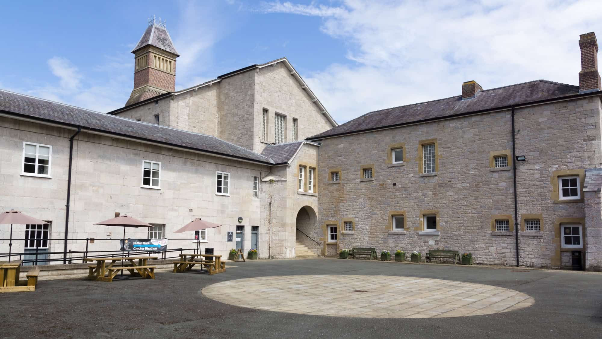 Courtyard of Ruthin Gaol, a historic stone prison in Wales, with pale brick buildings, barred windows, a central clock tower, and wooden picnic tables beneath umbrellas.
