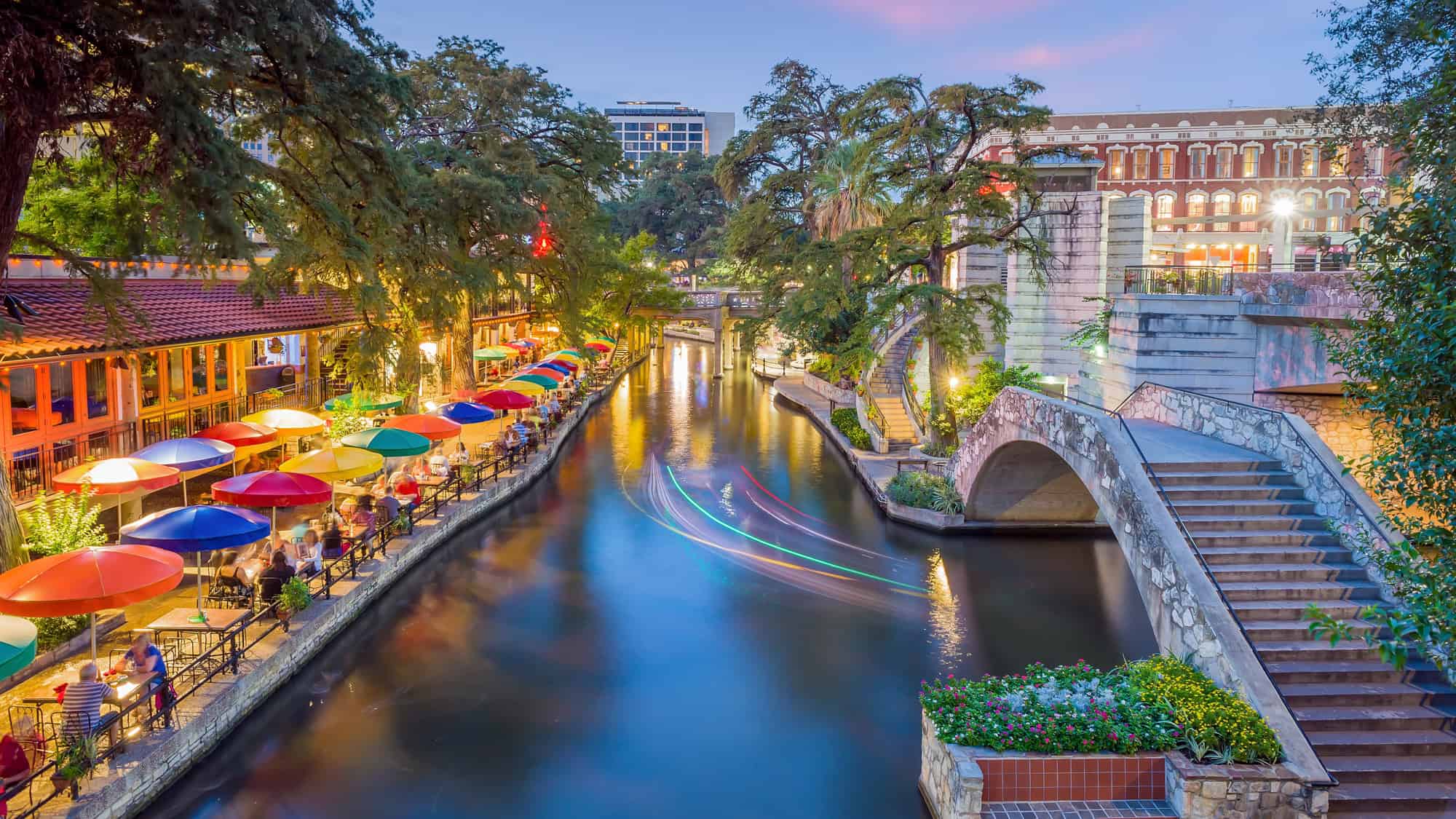A vibrant evening at the San Antonio River Walk, where colorful umbrellas line the riverside and restaurant-goers enjoy dinner beside the softly lit water.