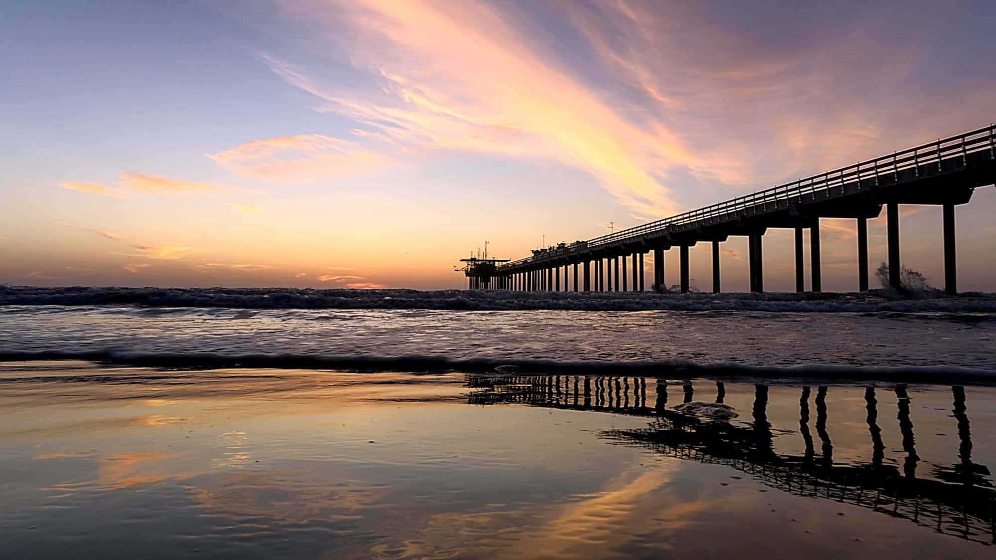 A tranquil shot of La Jolla Shores Pier in San Diego, California, stretching into the Pacific Ocean under pastel-streaked skies reflected in the wet sand.