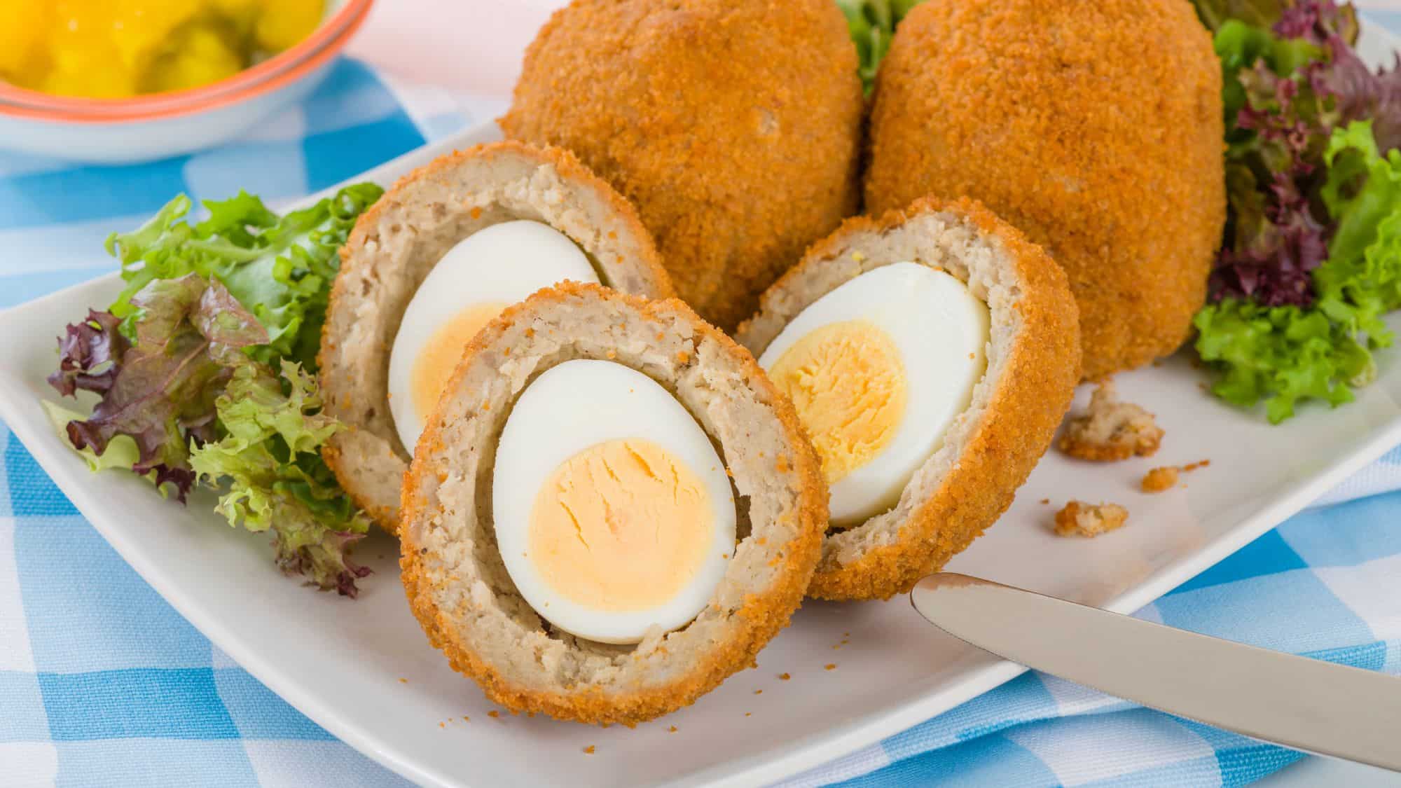A golden-brown, crispy-coated Scotch egg cut in half, revealing a hard-boiled egg encased in seasoned sausage meat, served with fresh greens on a white plate.