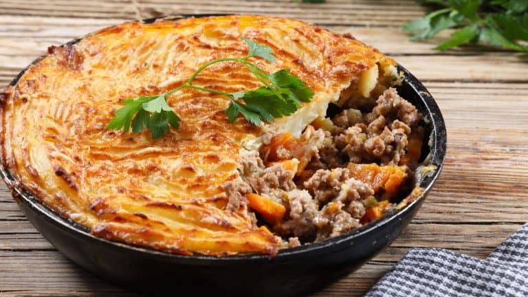 A traditional shepherd’s pie with a golden, crispy mashed potato topping, partially scooped to reveal a filling of minced meat, carrots, and peas, garnished with fresh parsley.