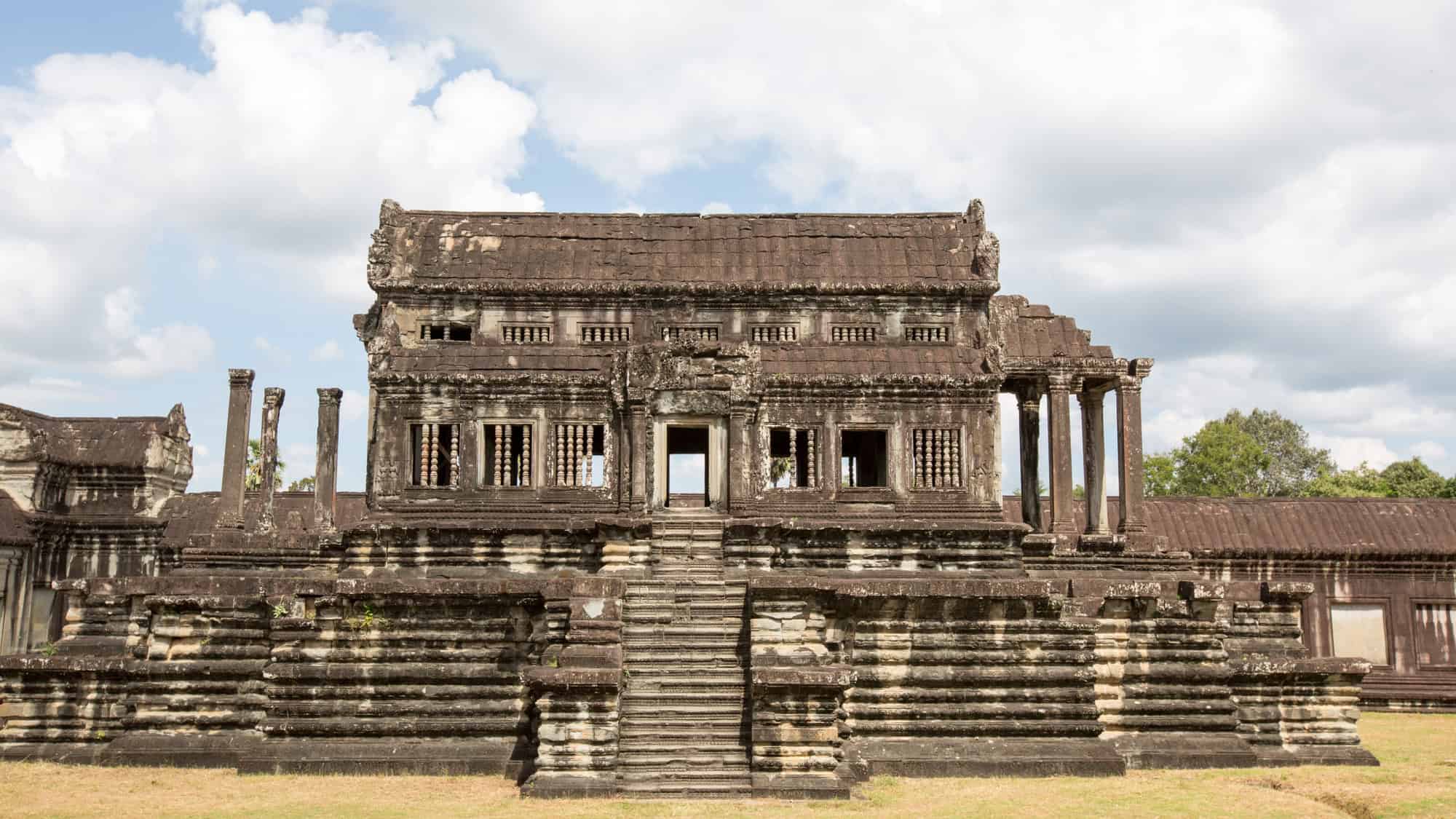 An ancient stone temple with intricate carvings and a central staircase, set against a partly cloudy sky and lush greenery.