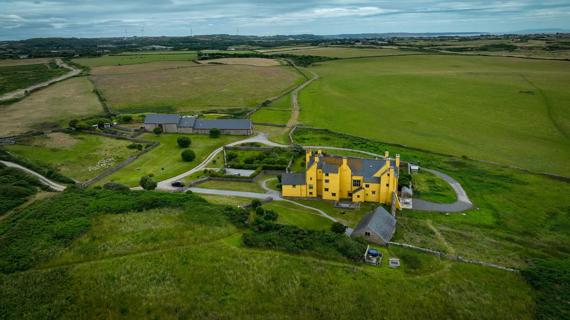 Aerial view of Sker House, a striking yellow historic manor surrounded by rolling green fields in Wales, with winding driveways and stone outbuildings dotting the property.