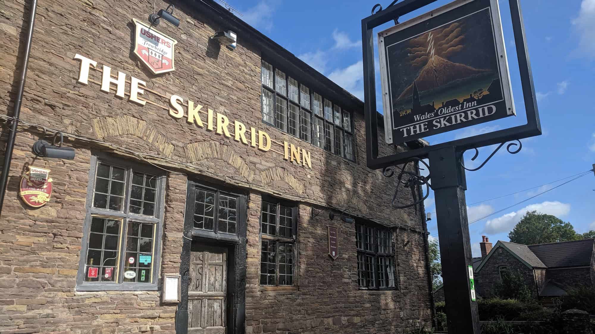 A historic stone building with leaded windows and a wooden door, The Skirrid Inn is marked by bold golden letters and signage claiming it as “Wales’ Oldest Inn.” A classic pub sign features a dramatic mountain scene with lightning, adding to its legendary reputation.