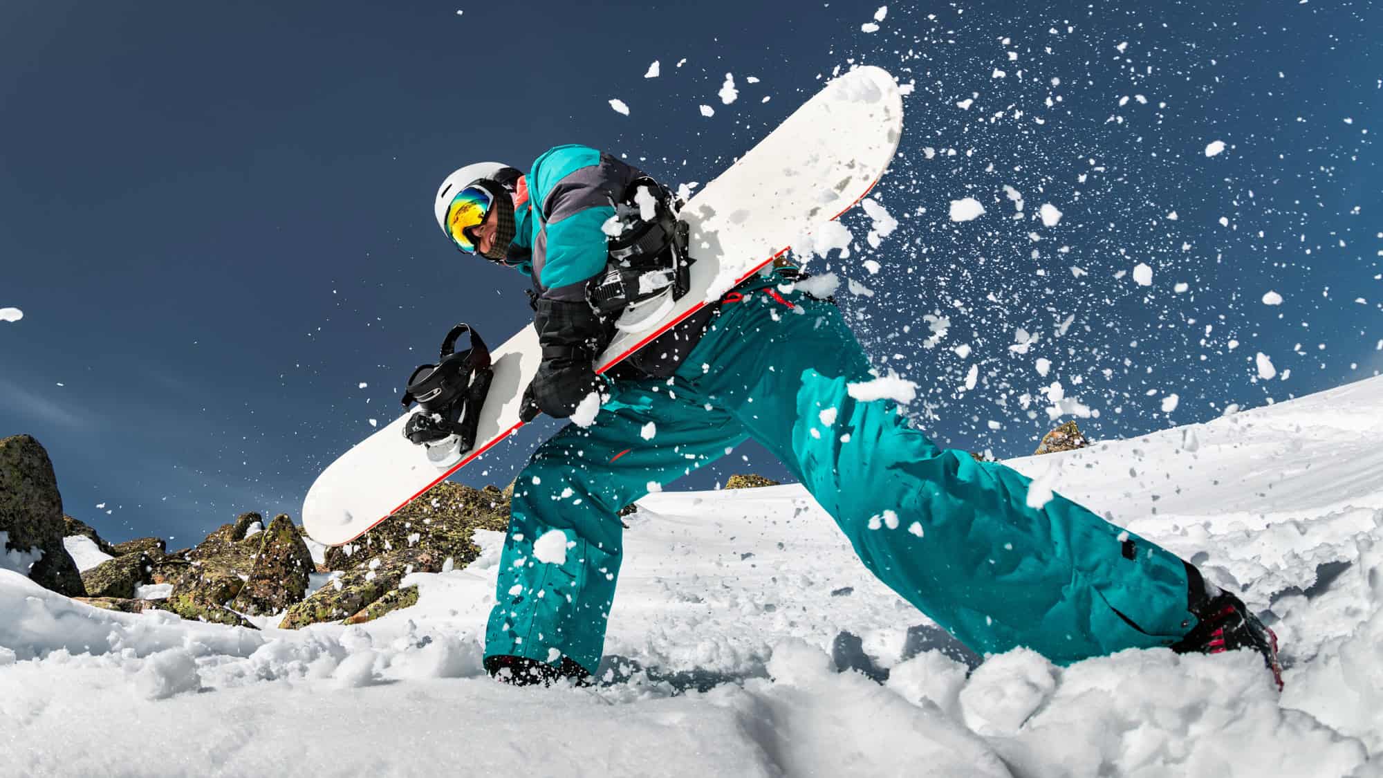 A snowboarder in teal snow gear, wearing a helmet and goggles, carries a white snowboard while kicking up fresh powder on a snowy mountain slope. The bright blue sky contrasts with the rugged, snow-covered terrain.