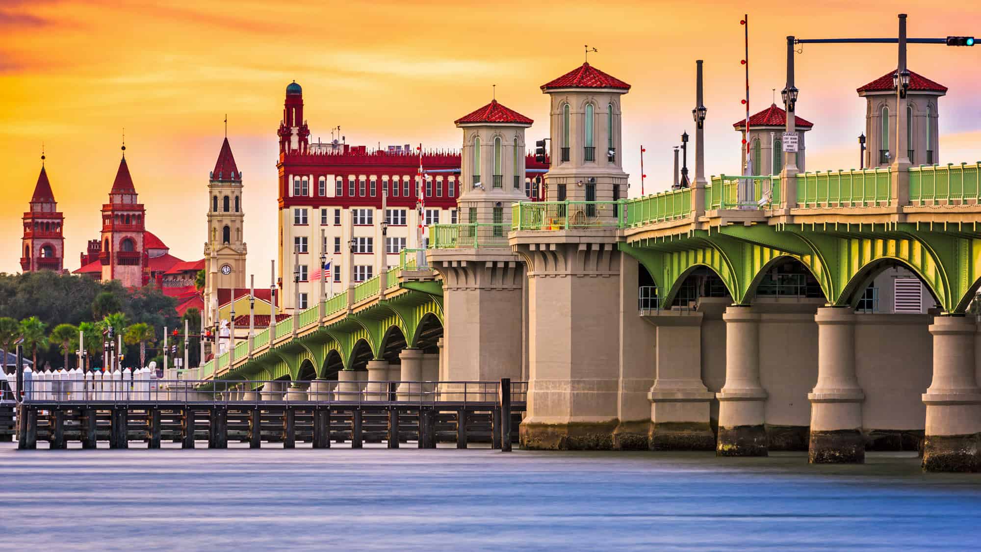 A vivid sunset casts a warm glow over the Bridge of Lions in St. Augustine, Florida, with the historic red-roofed towers of the city rising in the background.