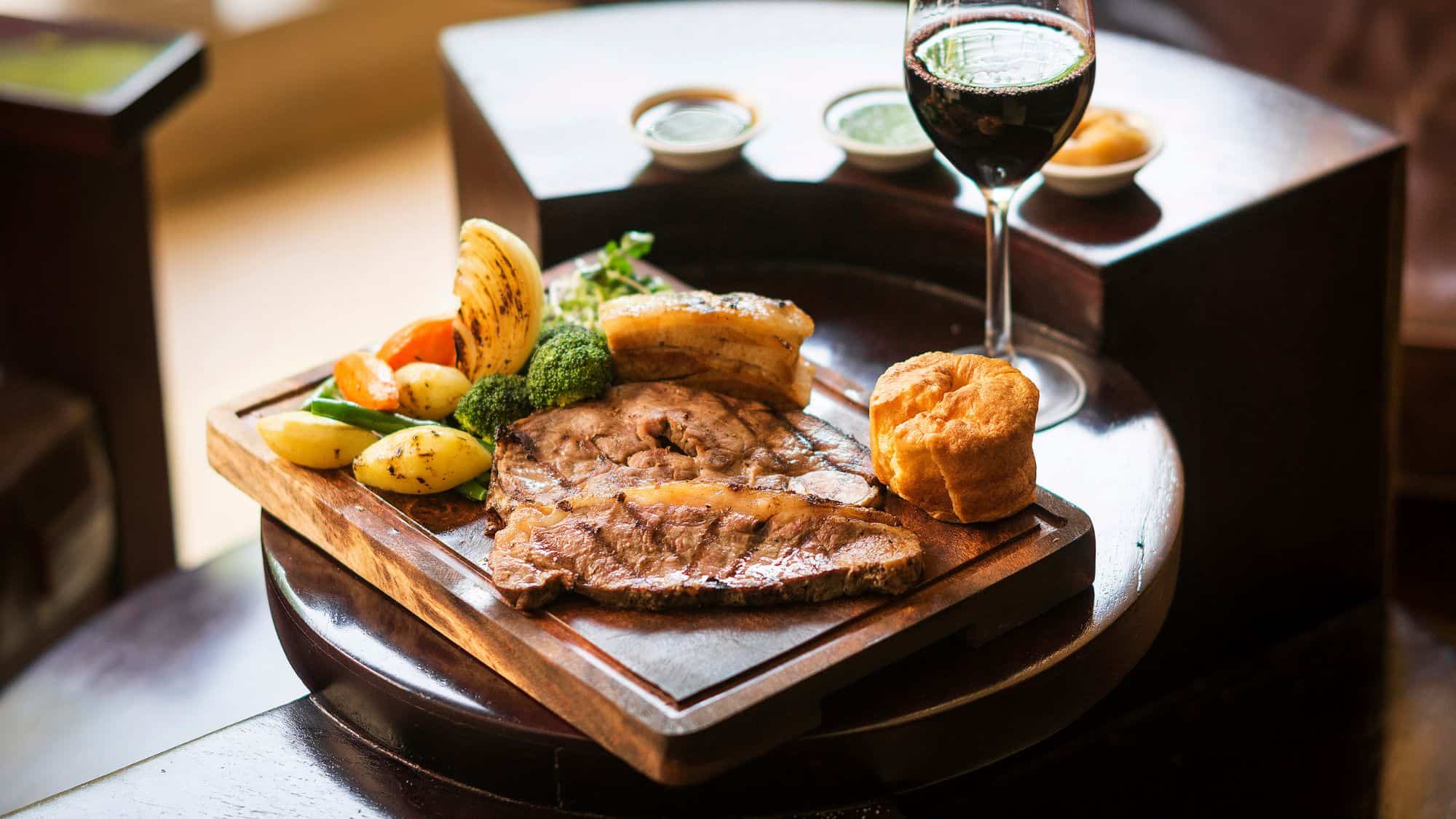 A traditional British roast dinner with a thick-cut piece of beef, Yorkshire pudding, roasted potatoes, grilled vegetables, and a glass of red wine on the side.