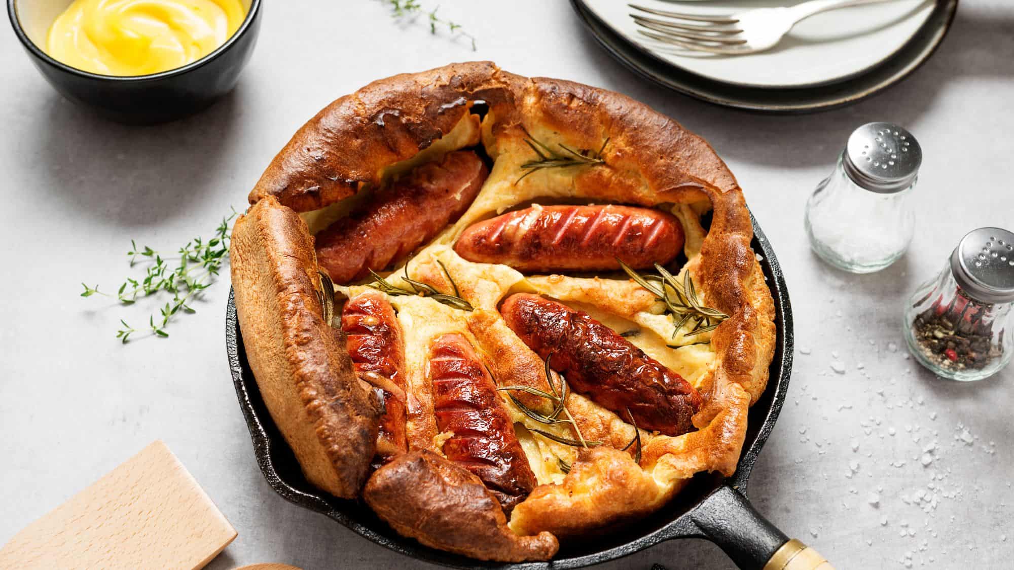 A skillet filled with sausages baked into a crisp, golden Yorkshire pudding batter, garnished with sprigs of rosemary, with a side of mustard and salt-and-pepper shakers.
