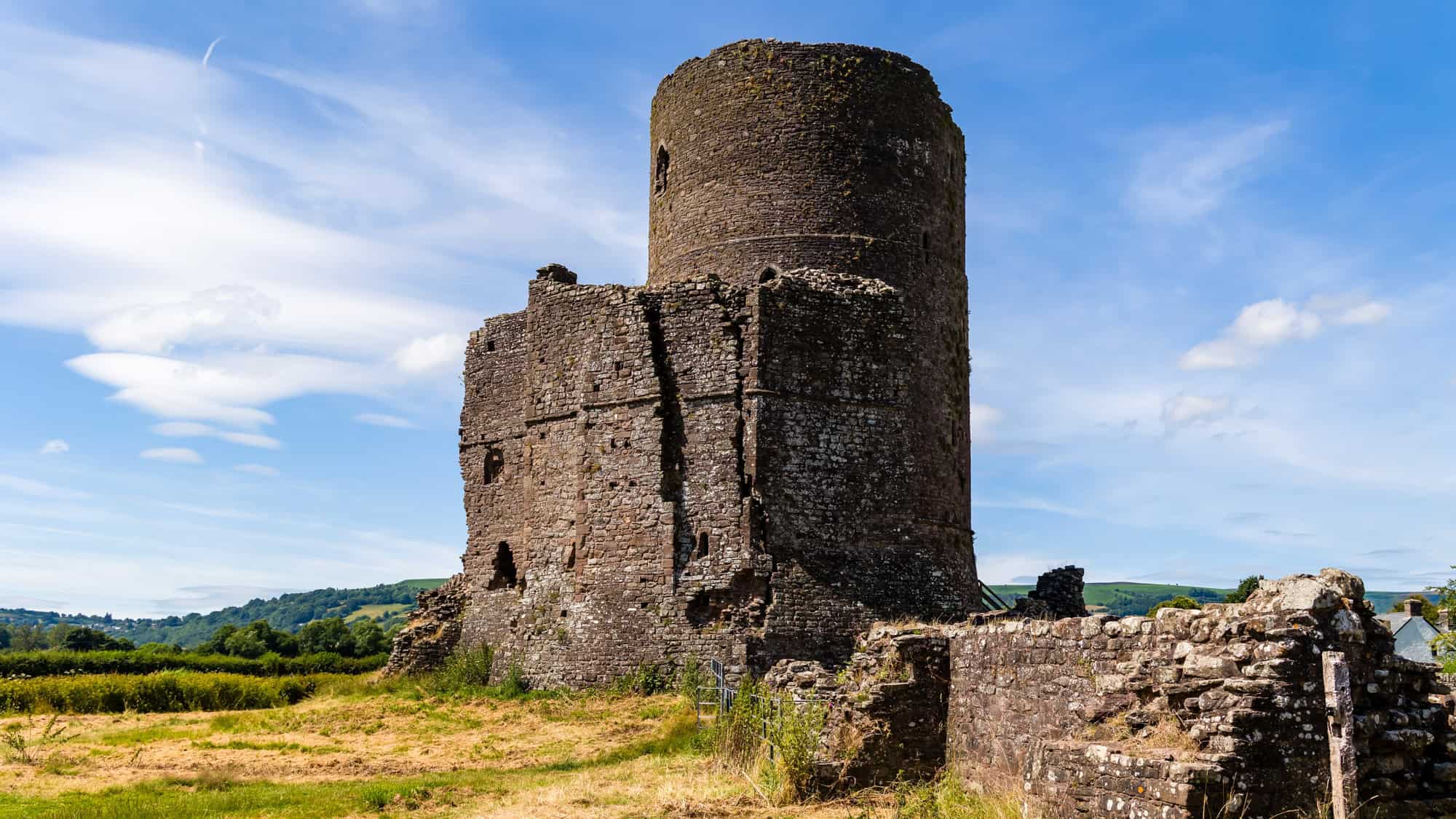Ruins of Tretower Court, showing a tall, cylindrical medieval tower and crumbling stone walls set in an open grassy field against a bright blue sky with wispy clouds.