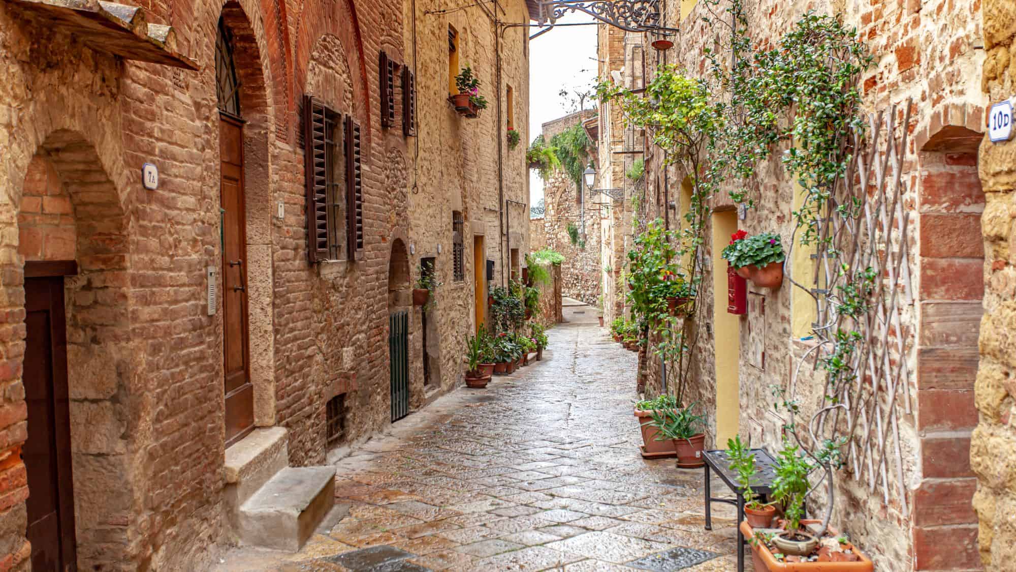 A charming cobblestone alley in Tuscany is lined with old brick buildings adorned with potted plants, green shutters, and arched doorways, evoking a rustic Italian ambiance.