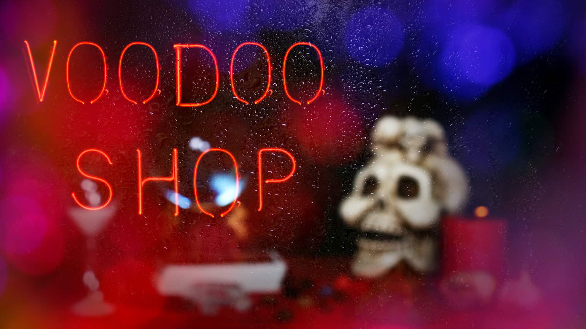 A glowing red neon sign reads "VOODOO SHOP" against a wet window with blurred colorful lights in the background. A skull decoration and ritual items are visible behind the glass.