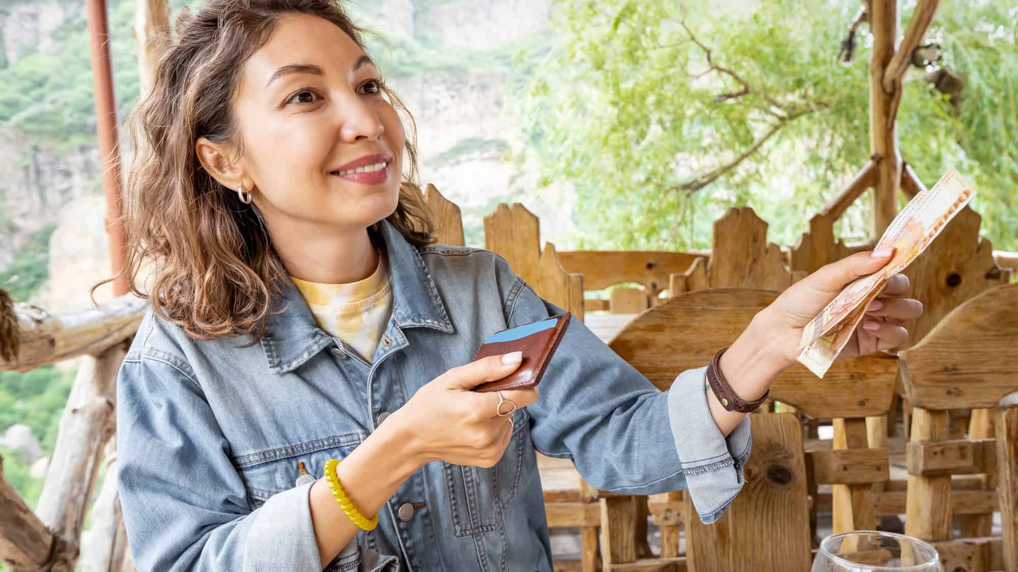 A smiling woman with curly light brown hair holds a cardholder and banknotes in an outdoor, rustic cafe setting with greenery and mountains in the background.
