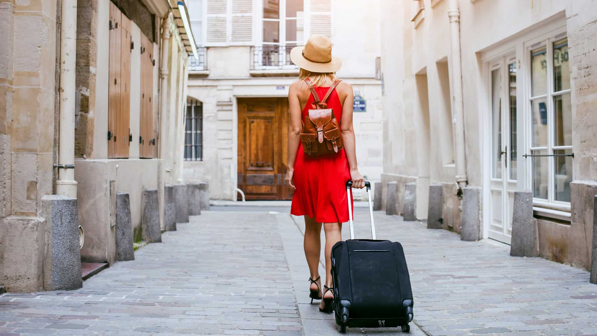 A stylish woman in a red dress and sun hat walks down a charming European alleyway, pulling a black rolling suitcase. The cobblestone street and classic architecture give the scene an old-world charm.