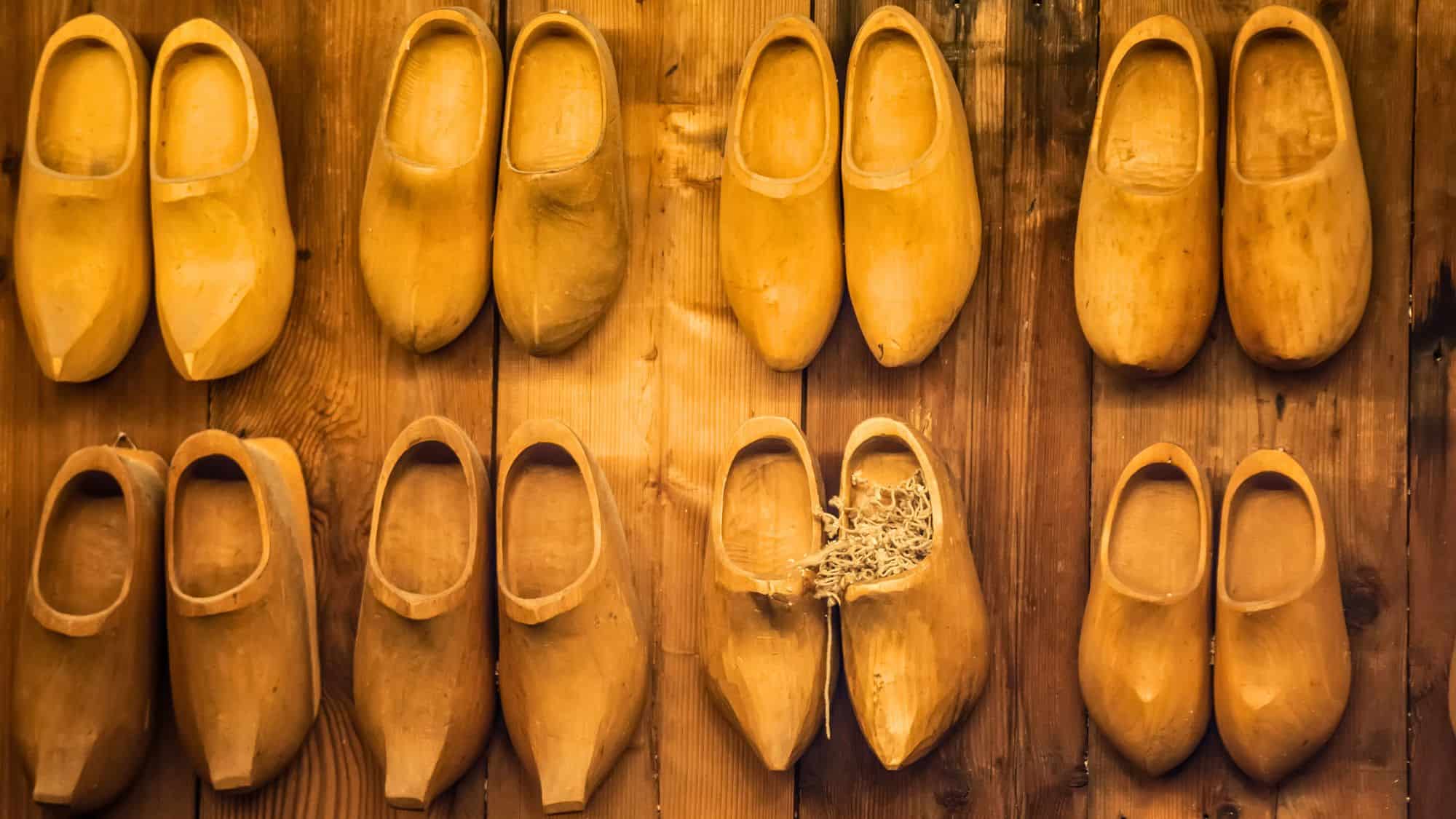 A collection of handcrafted wooden clogs in various shades of brown is displayed against a rustic wooden wall, with one pair showing signs of wear and straw inside.