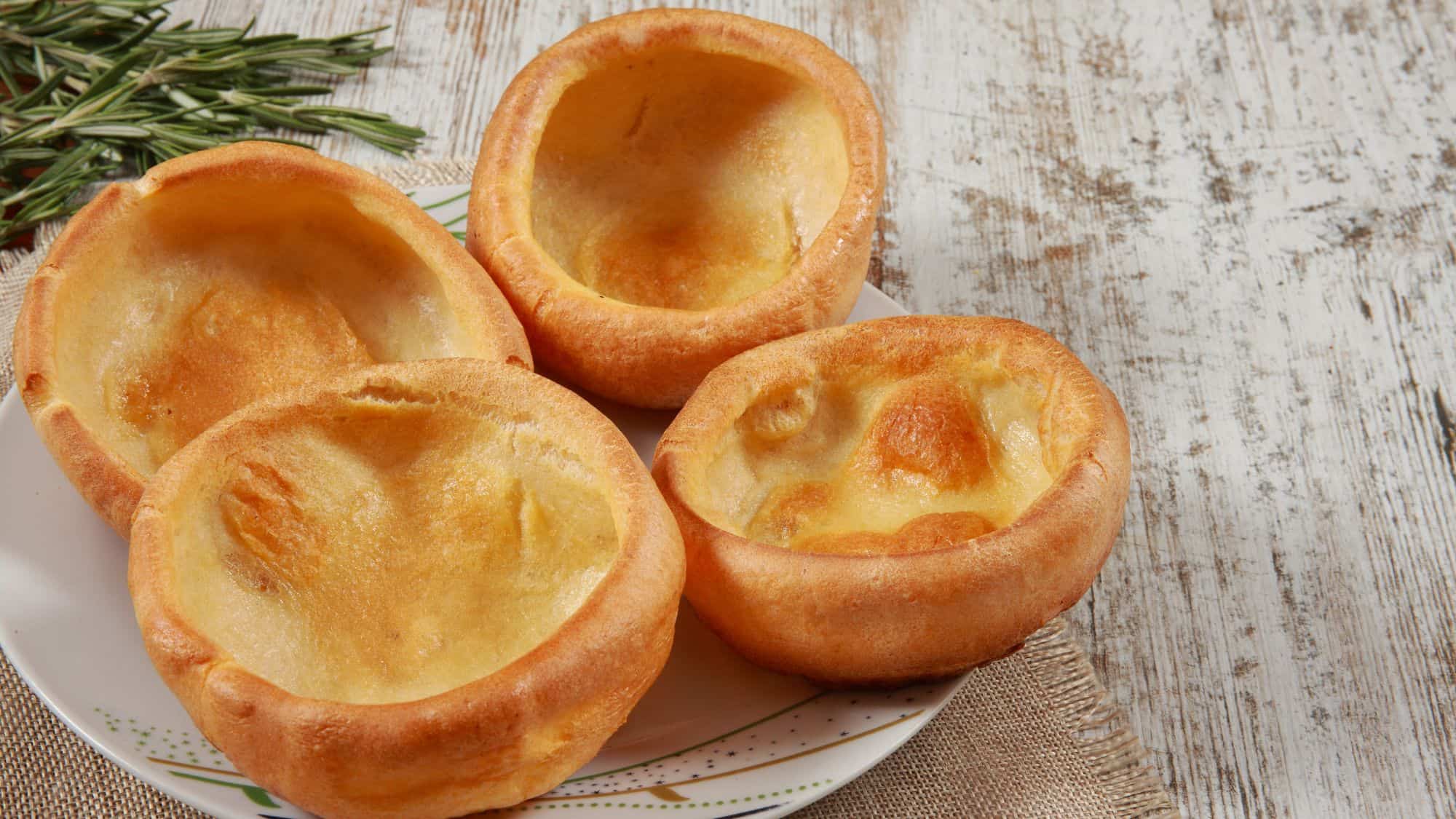 A plate of golden-brown Yorkshire puddings with a crisp, airy texture, set on a rustic wooden table with sprigs of fresh rosemary in the background.