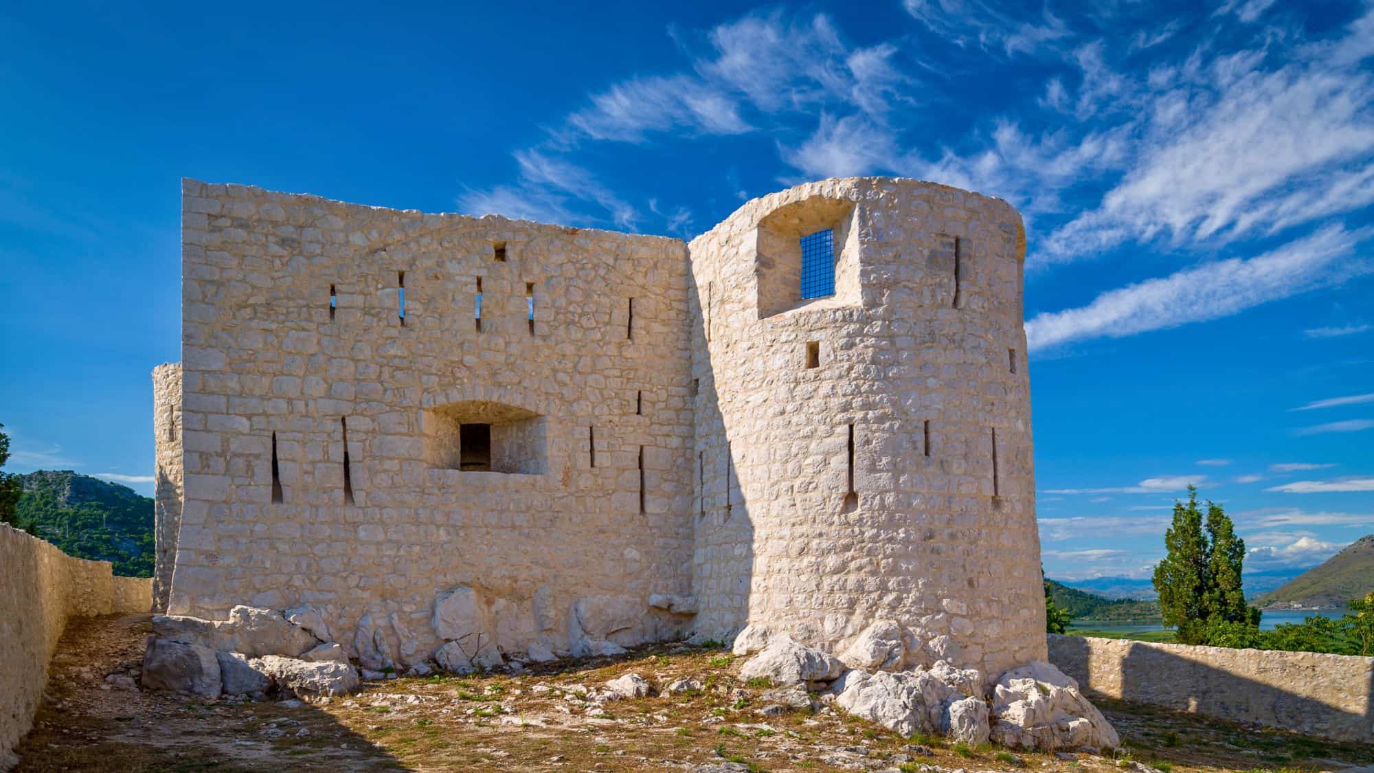 Brightly lit, well-preserved white stone walls of Besac Fortress in Montenegro, sitting atop rocky terrain under a vivid blue sky with wispy clouds.