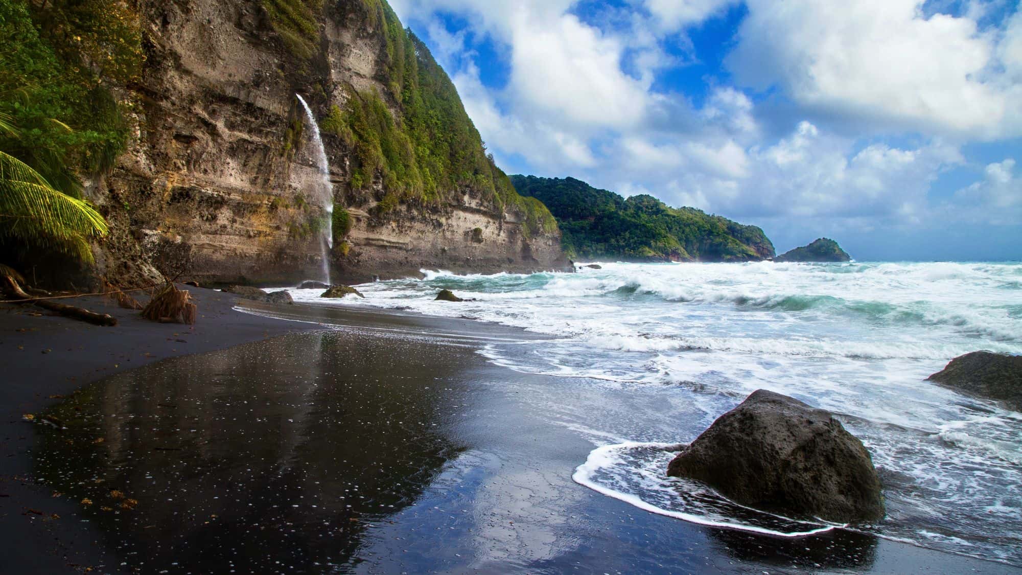 Dramatic view of a black sand beach with foamy waves crashing against the shore, a tall waterfall cascading down a cliffside, and lush green hills rising in the distance under a partly cloudy sky.