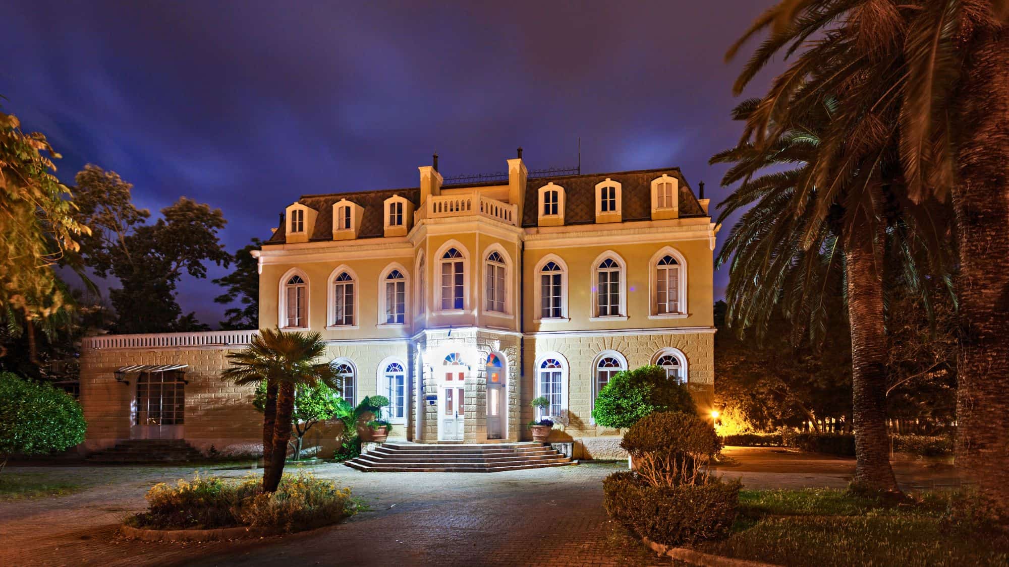 Elegant night view of the Castle of King Nikola in Bar, Montenegro, with its symmetrical façade lit warmly, surrounded by palm trees and manicured greenery.