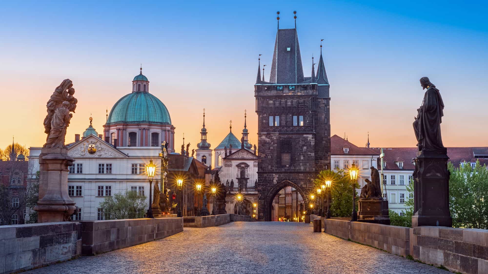 An iconic view of Prague’s Charles Bridge at sunset, with vintage-style lampposts, baroque statues, and the Old Town Bridge Tower silhouetted against a colorful sky.