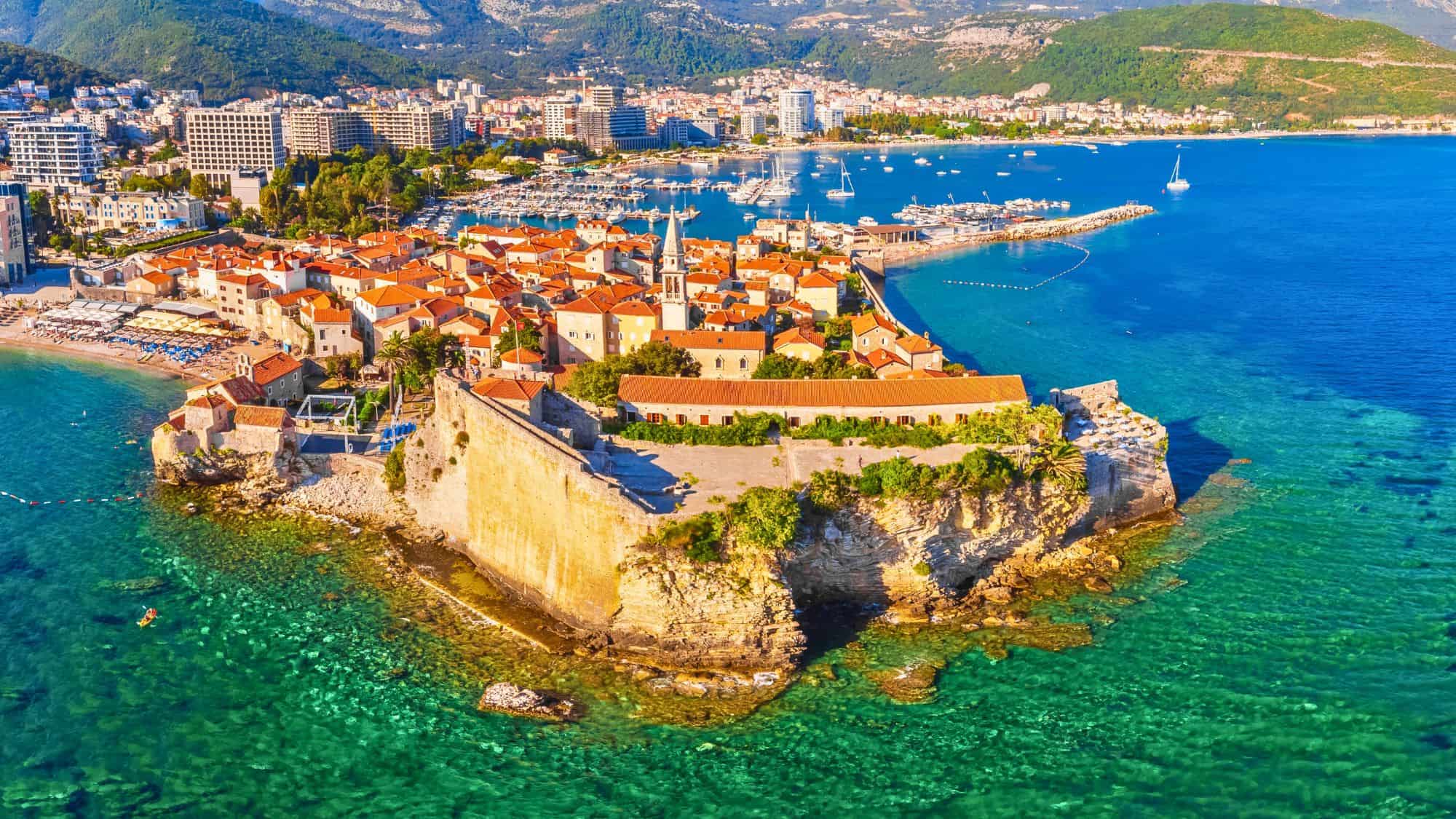 Aerial view of Budva's seaside citadel in Montenegro, with terracotta rooftops, sandy beaches, and vibrant turquoise waters contrasting the rocky coastline.