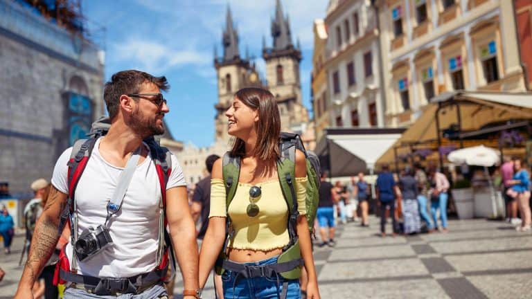 A happy couple with backpacks and a camera stroll through Prague’s Old Town Square, smiling at each other with the spires of the Church of Our Lady before Týn in the background.