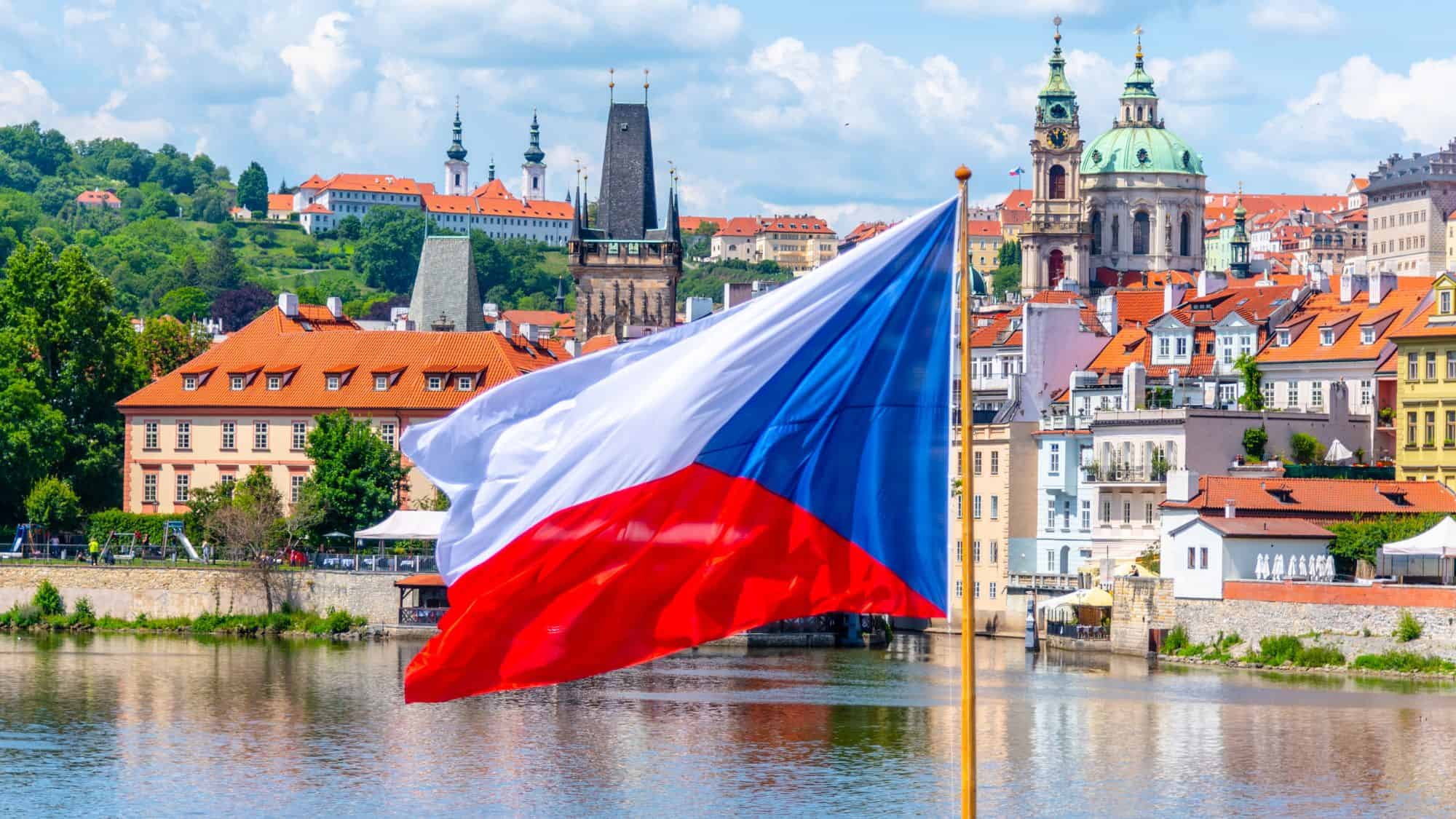 The Czech Republic flag waves proudly in the foreground with a view of the Vltava River, pastel-colored buildings, and historic church towers in the scenic Prague skyline.