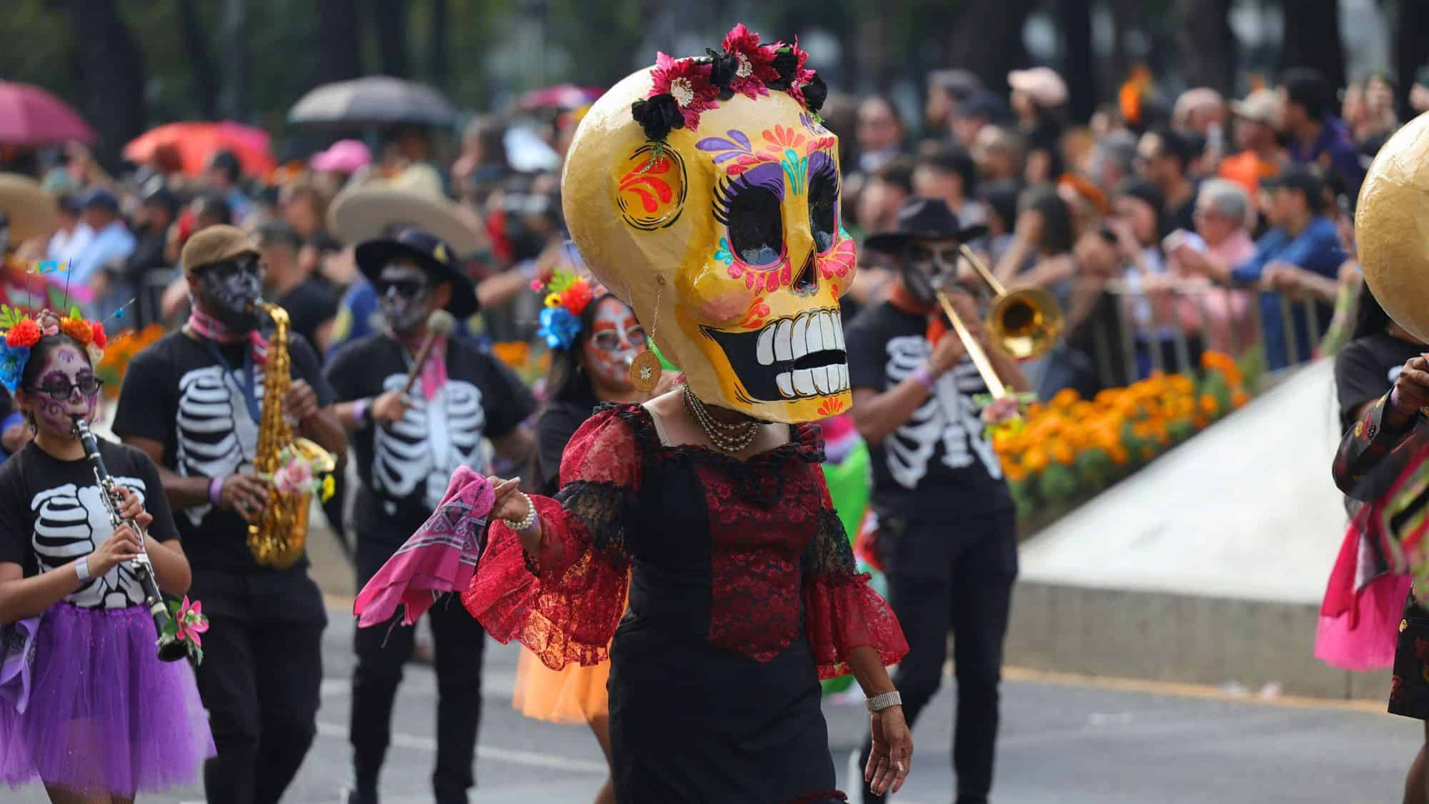 A vibrant Day of the Dead parade with participants in skeleton costumes and colorful face paint, including a person wearing a large decorative skull mask and red lace dress leading a lively street procession.