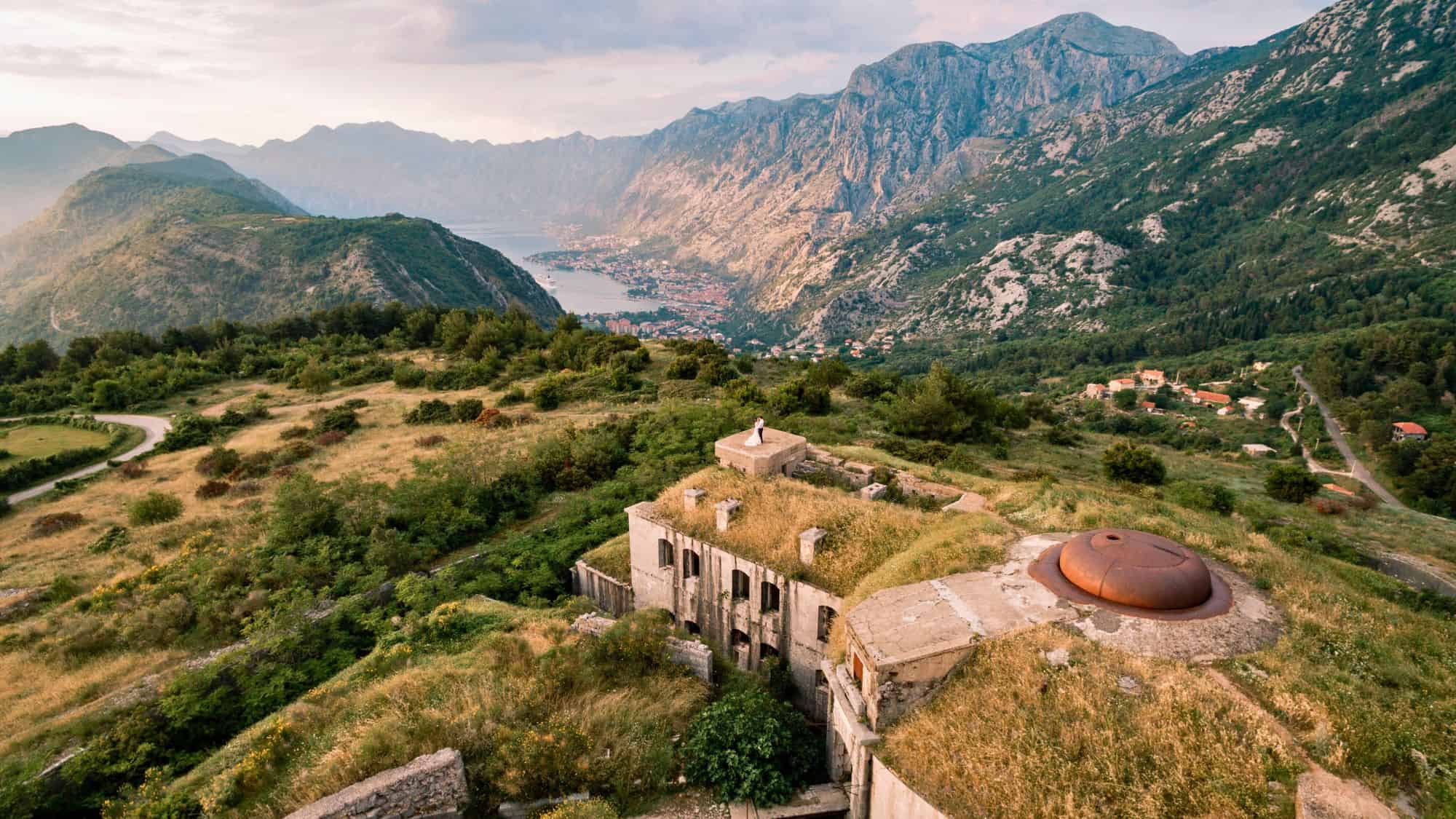A panoramic aerial view of Fort Gorazda, a decaying mountaintop fortress overgrown with grass, with stunning views of the Bay of Kotor and dramatic mountain ridges stretching into the distance. A couple stands atop one of the concrete bunkers, emphasizing the site's vastness and beauty.