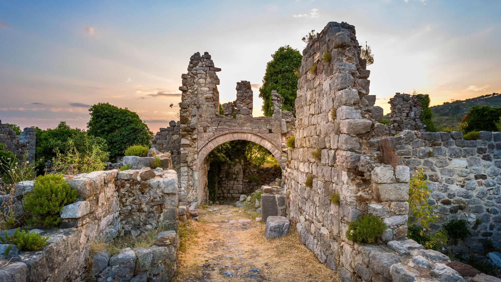 Crumbled stone walls and archways of the ancient Fortress of Stari Bar, Montenegro, backlit by a pastel-toned sky at sunset with overgrown vegetation.