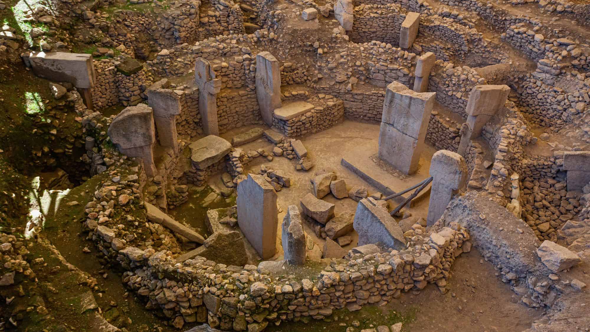 Overhead view of the ancient stone circle ruins at Göbekli Tepe in Turkey, featuring massive T-shaped pillars arranged in circular enclosures surrounded by rocky excavated earth.