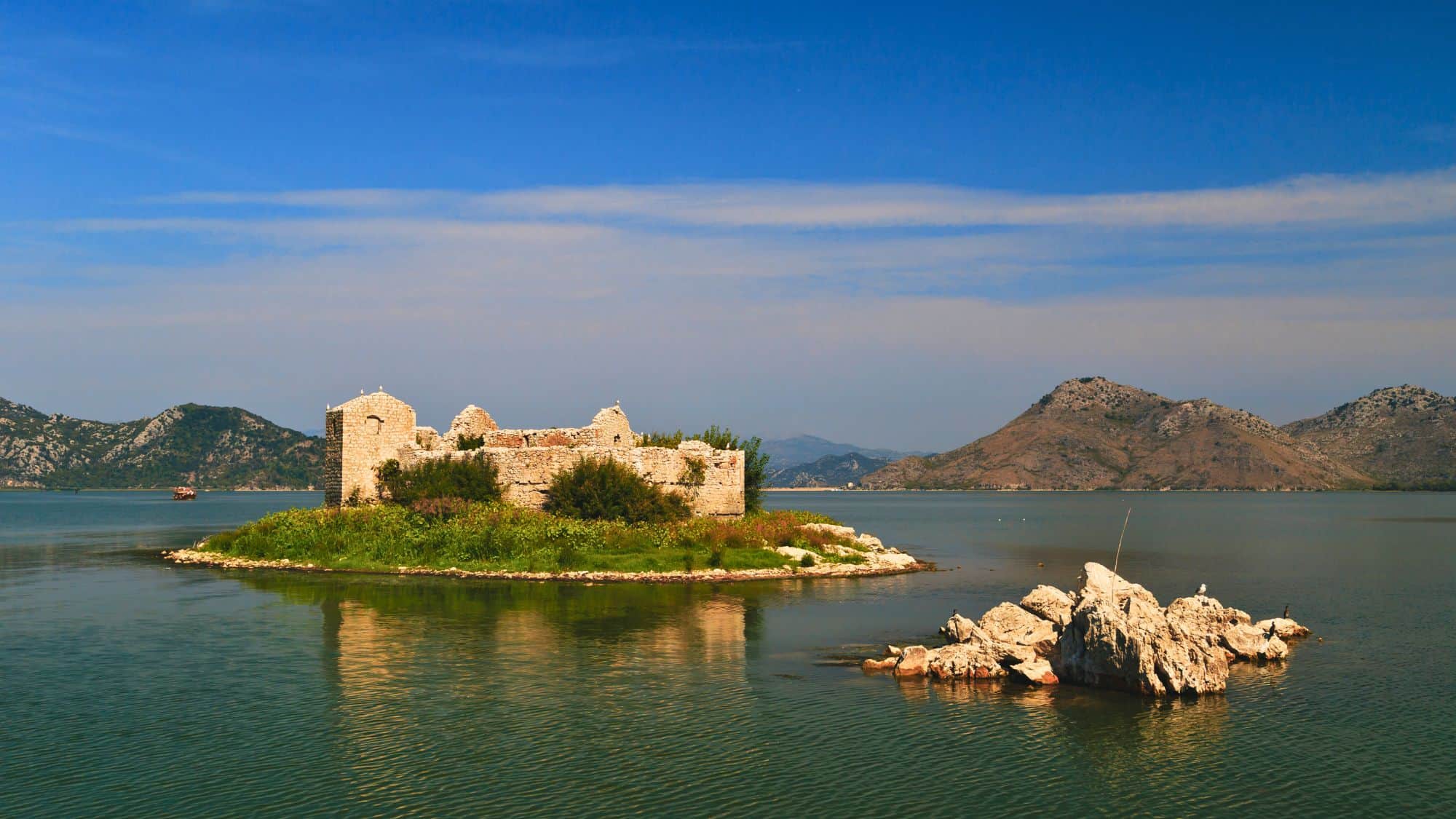A small, rugged island in Lake Skadar hosts the crumbling stone remains of Grmožur Fortress, surrounded by still water and flanked by distant, hazy mountains. Wild greenery has partially overtaken the old prison walls, blending it into the serene natural surroundings.